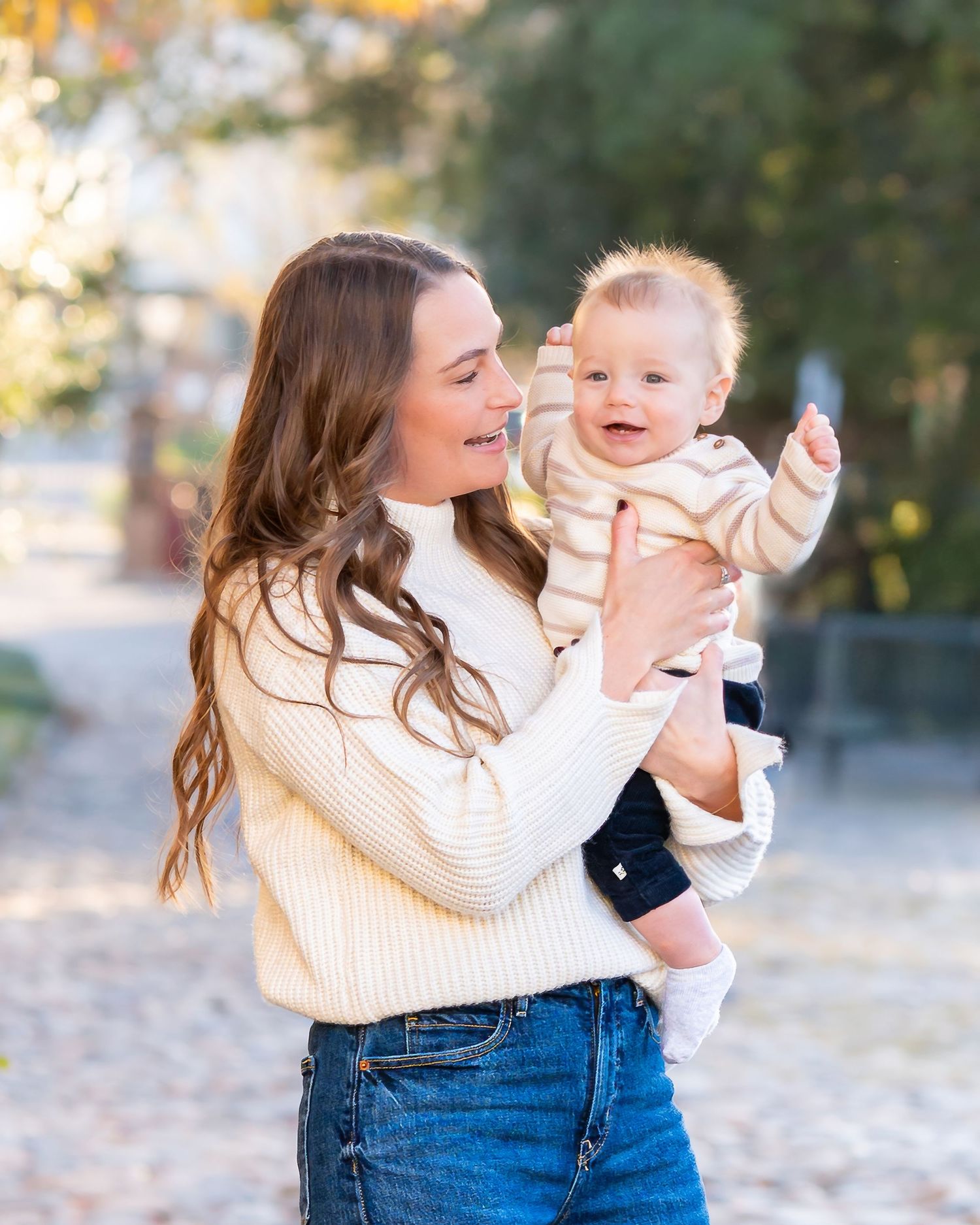 Parent and baby share playful moments in cream sweater against autumn outdoor backdrop.