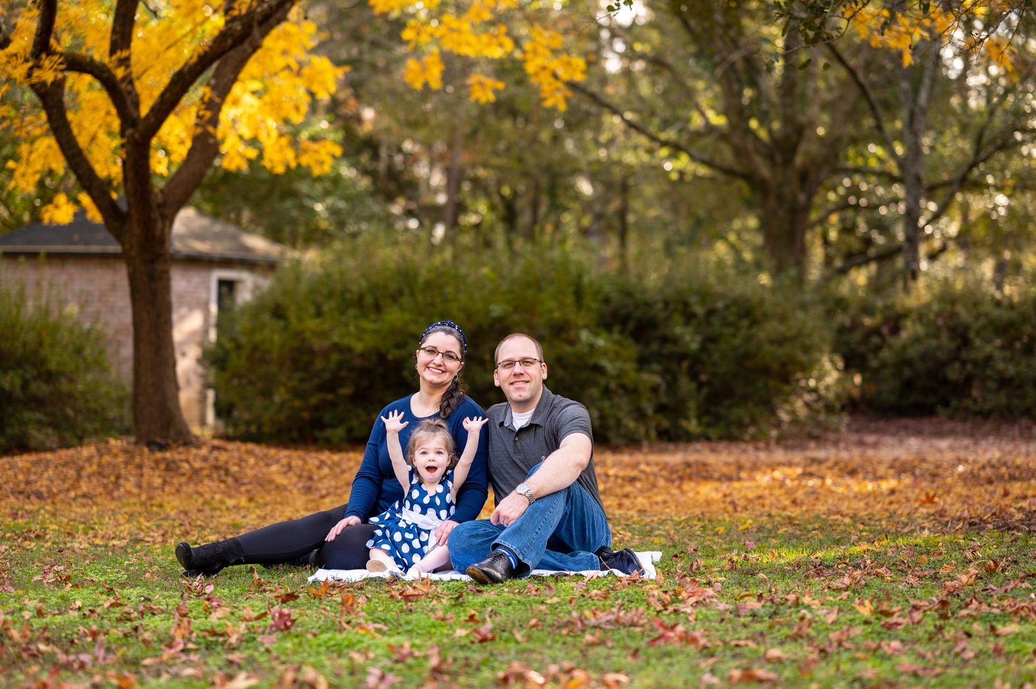 Amanda and Tyler sit with their toddler daughter at a park surrounded by fall foliage.