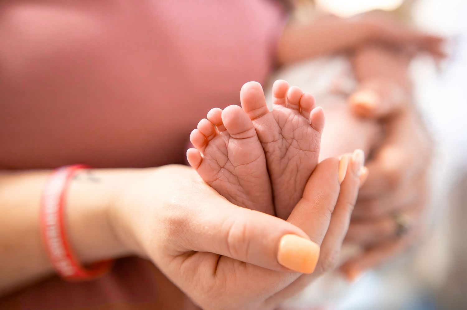 Adult hands gently holding a newborn baby's tiny feet against a soft blurred background.