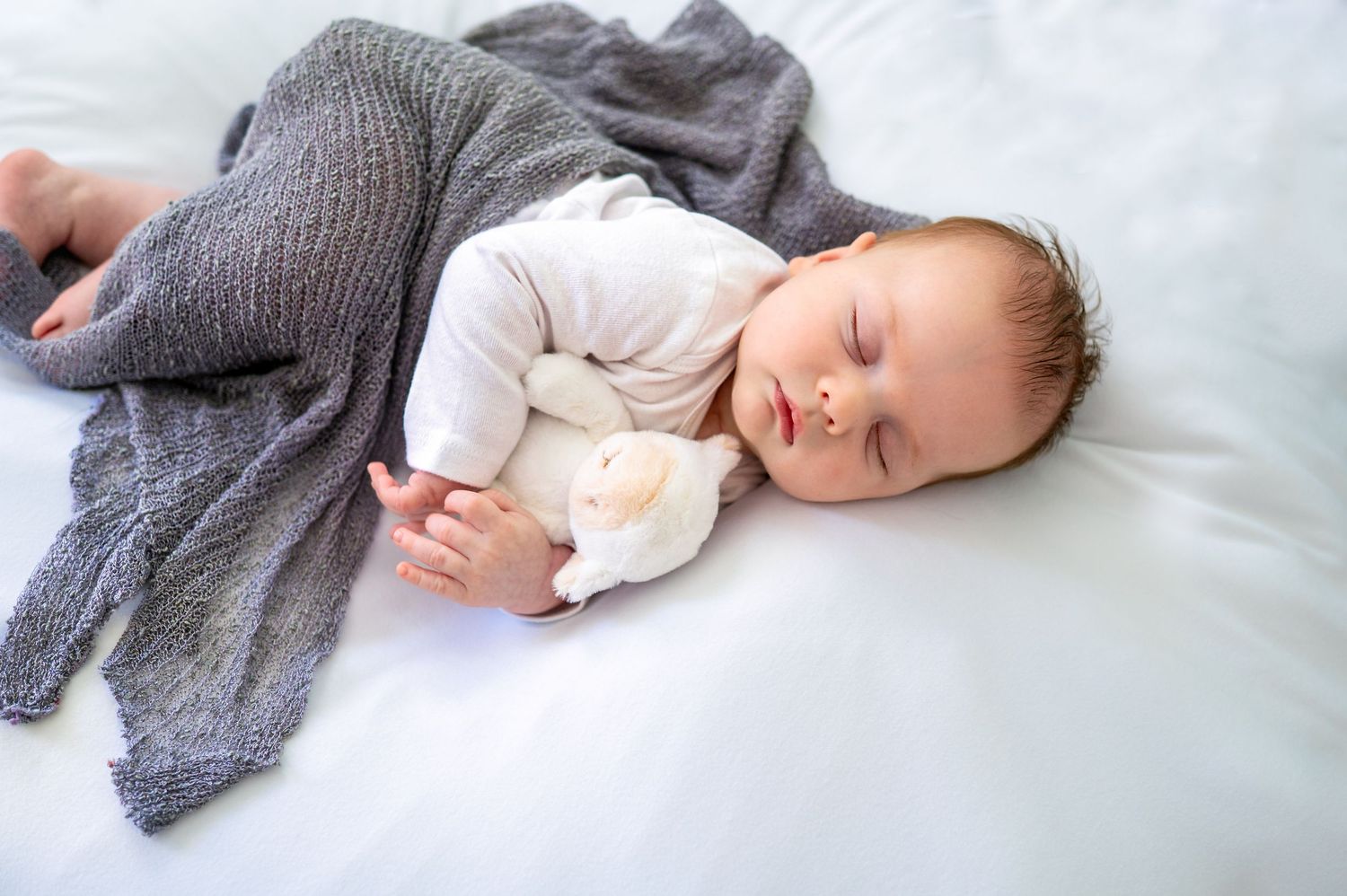 Peaceful newborn baby sleeping on white bedding, wrapped in gray knit blanket, cuddling a small white stuffed toy.