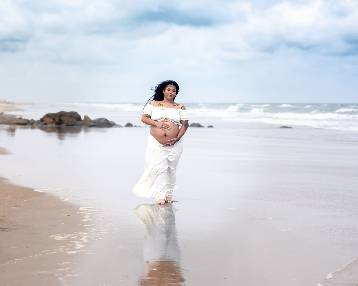 Pregnant woman in white dress walking on beach shoreline with waves and rocks in background.