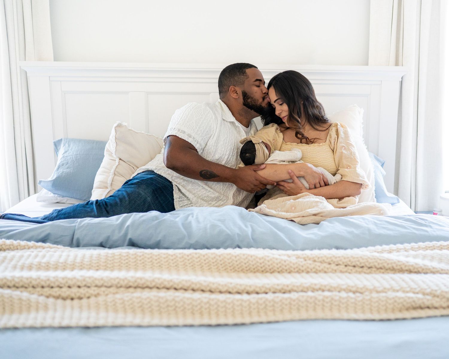 Couple sharing a tender kiss on a bed while cradling their newborn baby wrapped in a blanket.