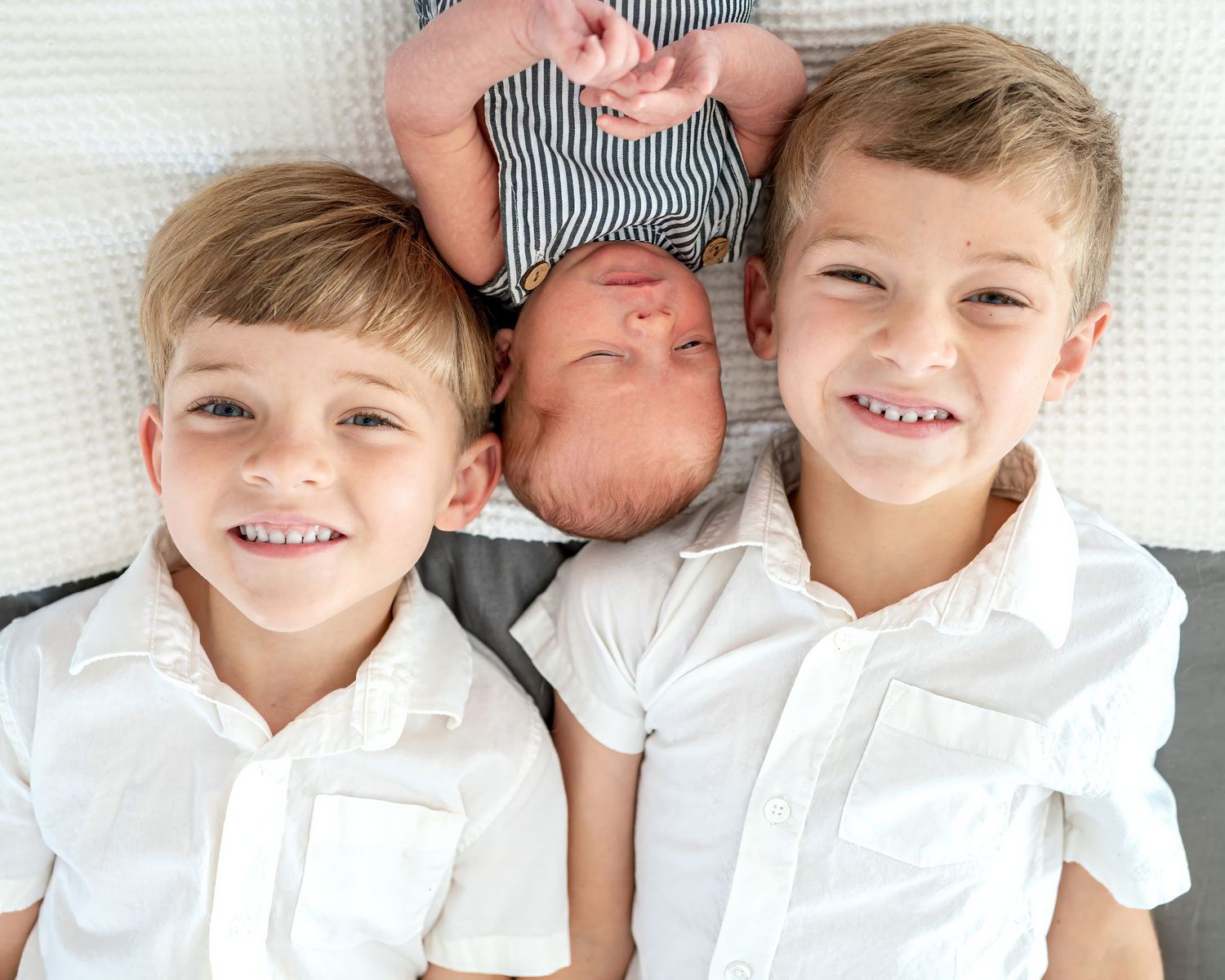 Children in white shirts pose with newborn sibling on white bedding.