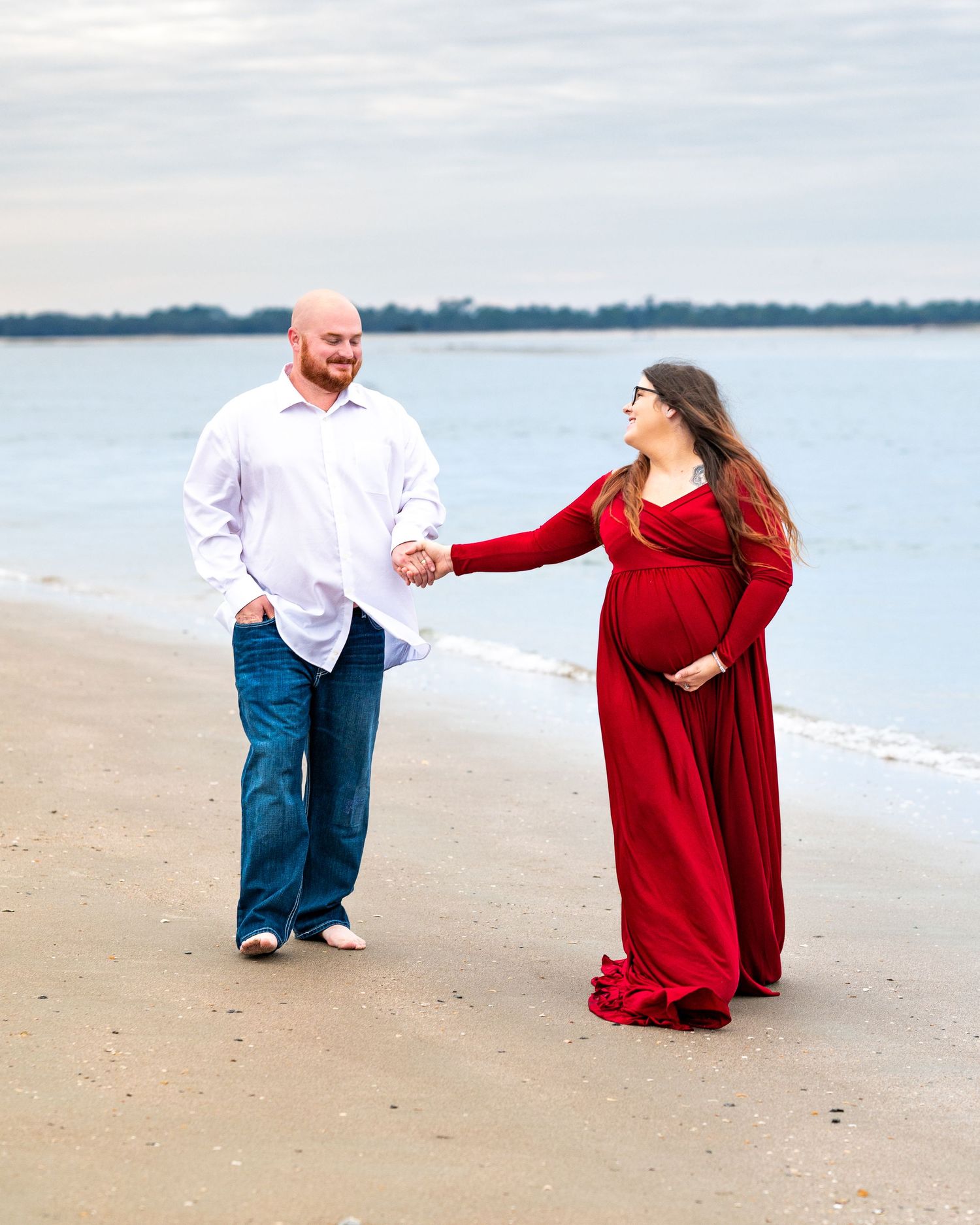 Couple walking hand in hand on a beach at sunset, with the woman wearing a flowing red maternity dress.