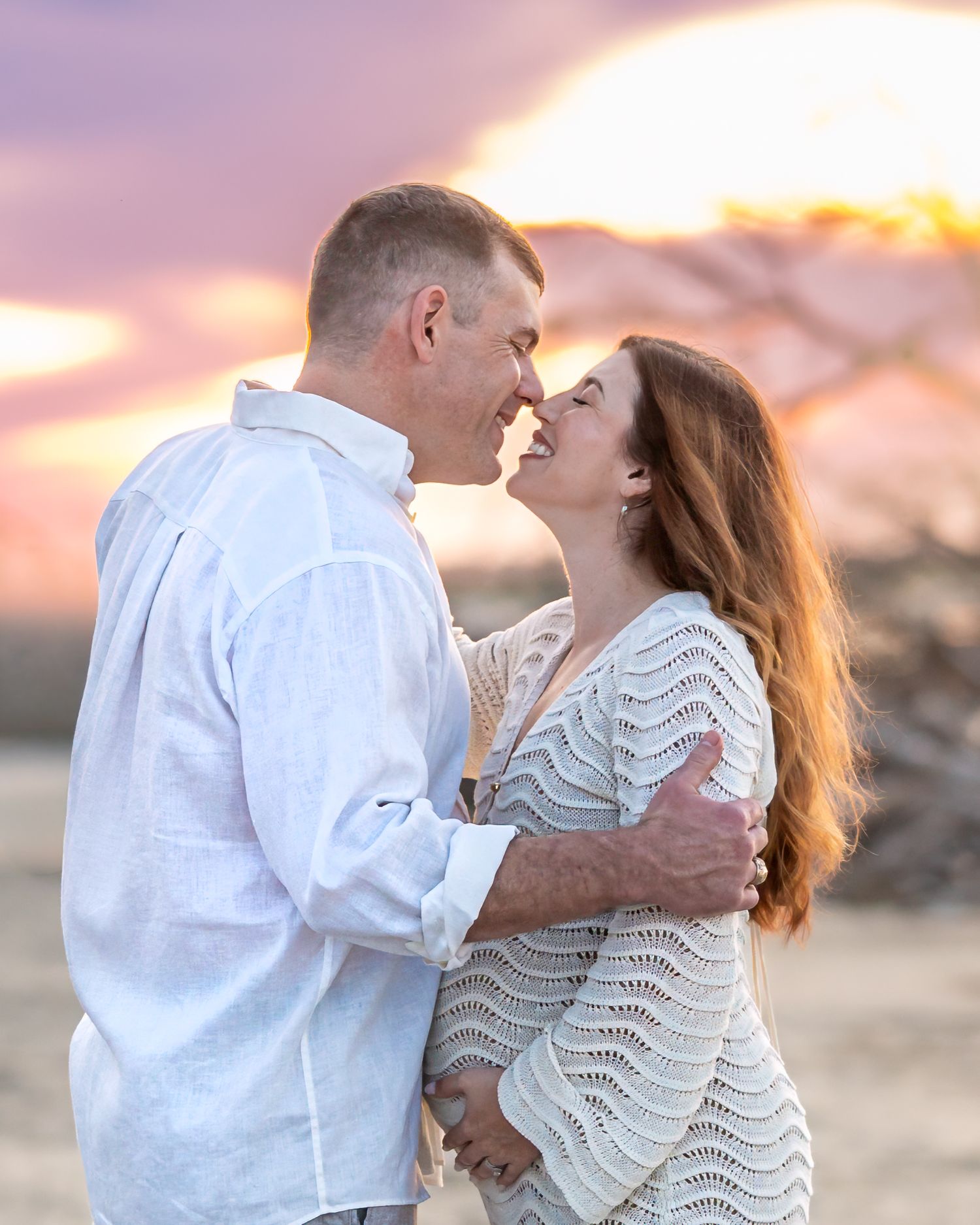 Romantic couple sharing an intimate moment at sunset on a beach, dressed in white and cream outfits during their maternity session.