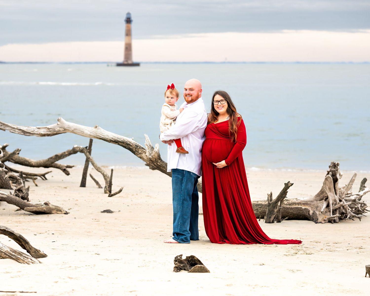 Family portrait on beach with driftwood and lighthouse in background during sunset, featuring flowing red dress.