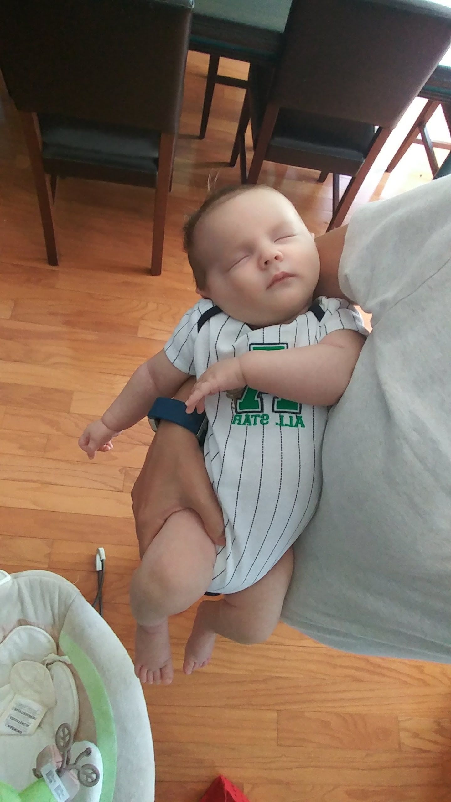 A baby wearing a striped onesie sleeps peacefully in his mother's arm with hardwood flooring visible in the background.