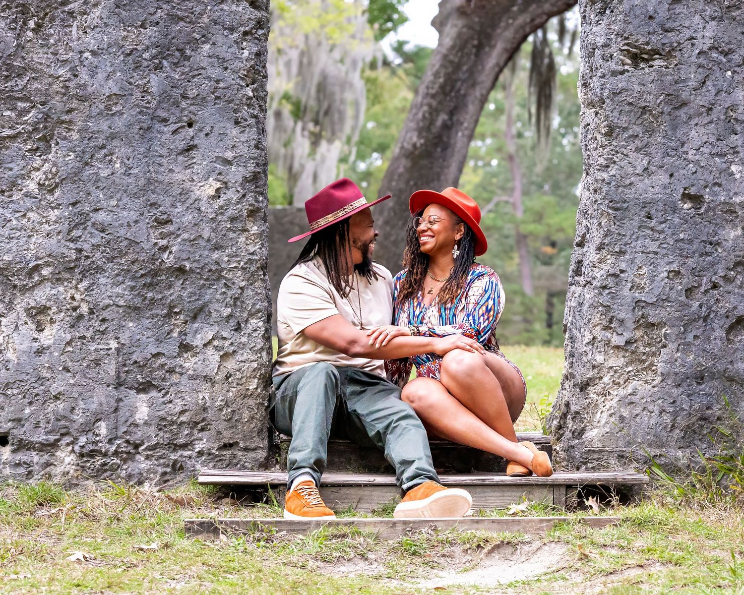 Couple sitting by rustic stone wall enjoying playful moments in an outdoor setting.