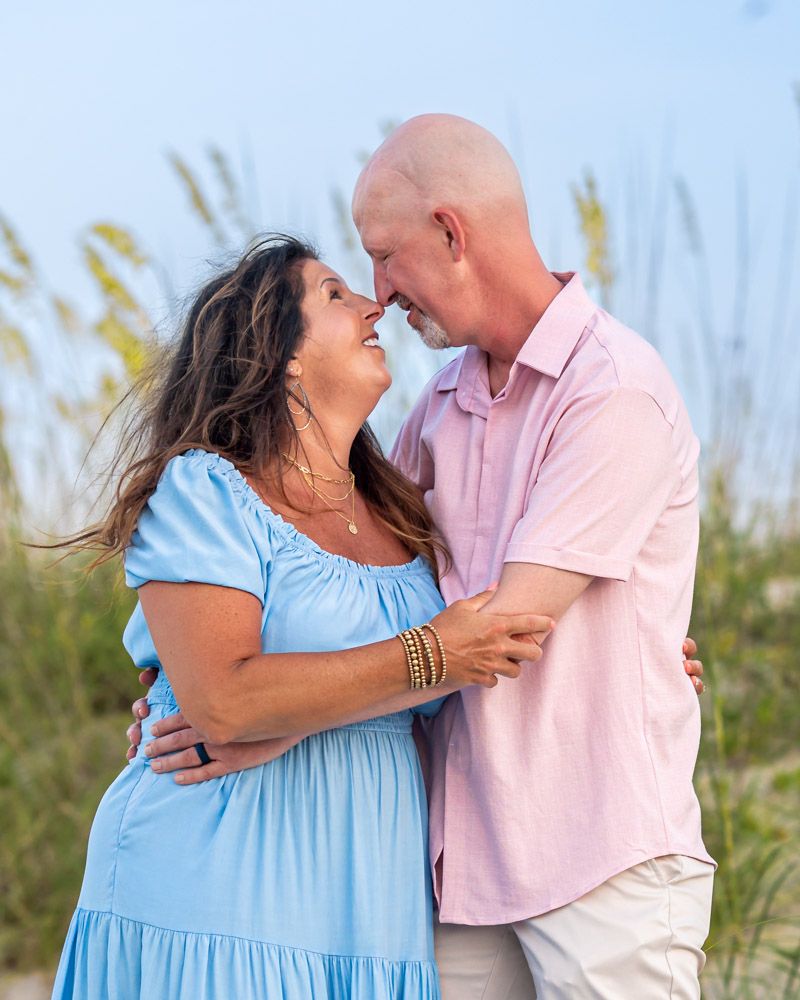 A couple shares a romantic moment on a beach, with the woman in a blue dress and man in a pink shirt touching foreheads.