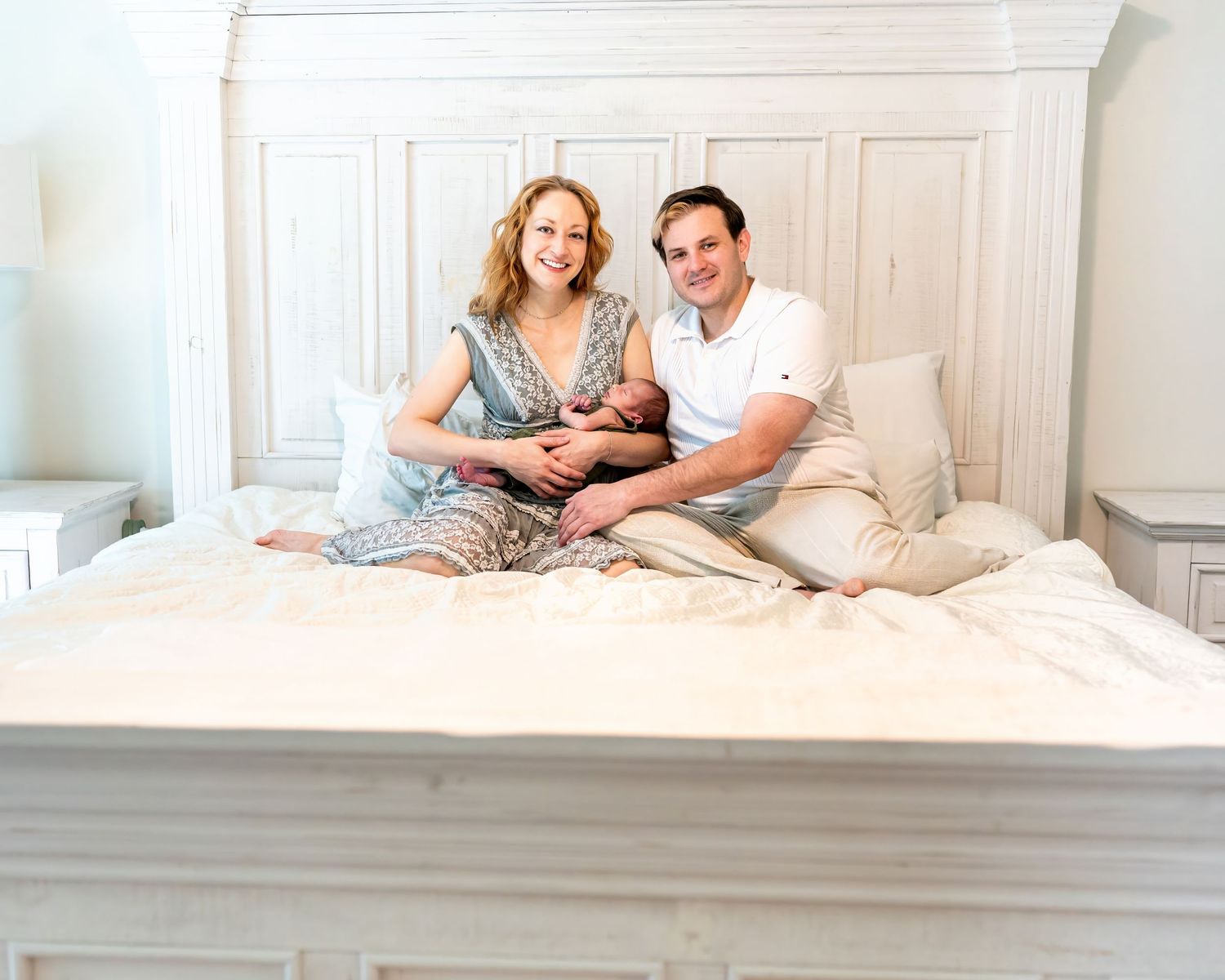 A happy couple sits on a white bed holding their newborn baby in a bright, elegantly decorated bedroom.