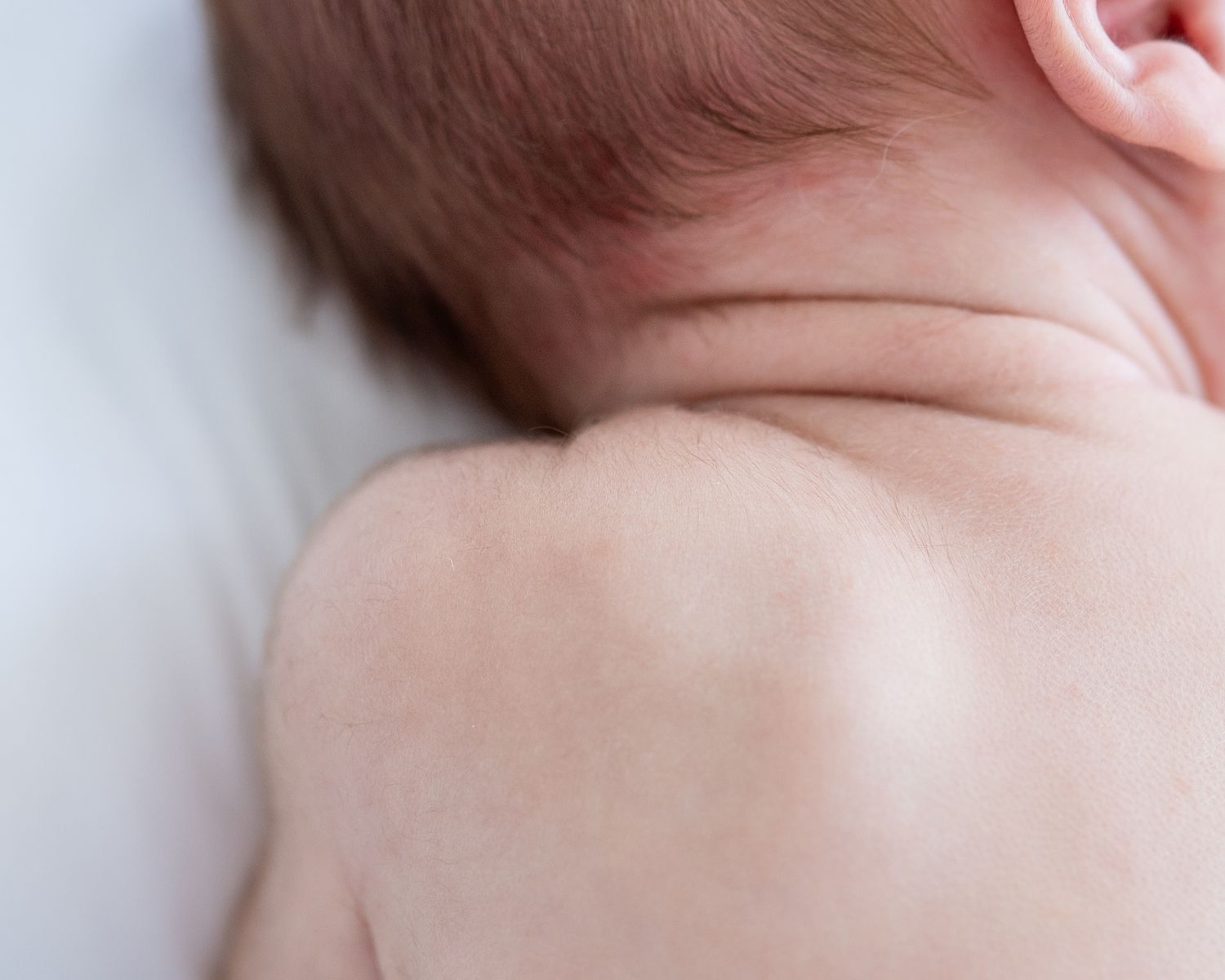 Close-up of a newborn baby lying on a white surface, showing delicate skin on the neck, shoulder, and back.