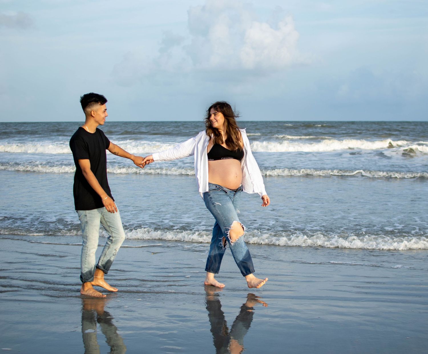 Alice and Brent walk along the ocean shore holding hands during their family maternity session