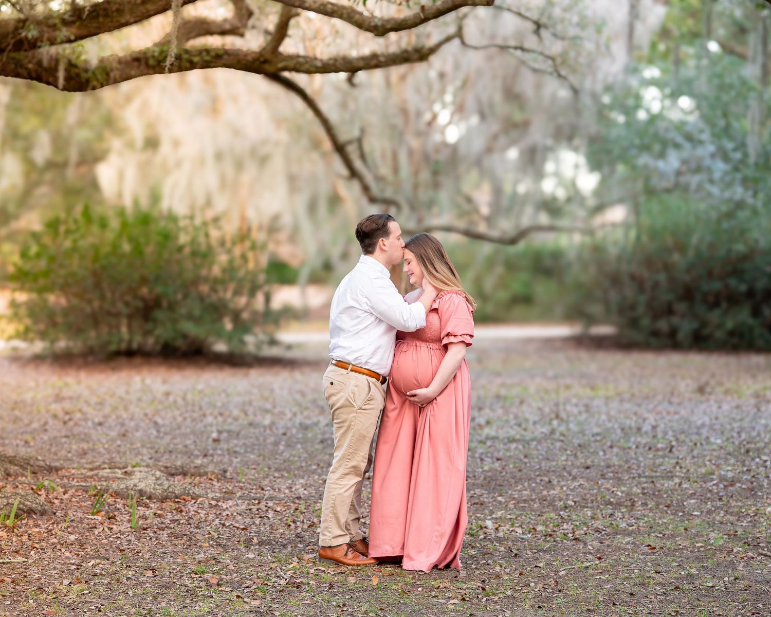 A romantic couple stands beneath Spanish moss draped trees in a park wearing pink dress and khaki attire.