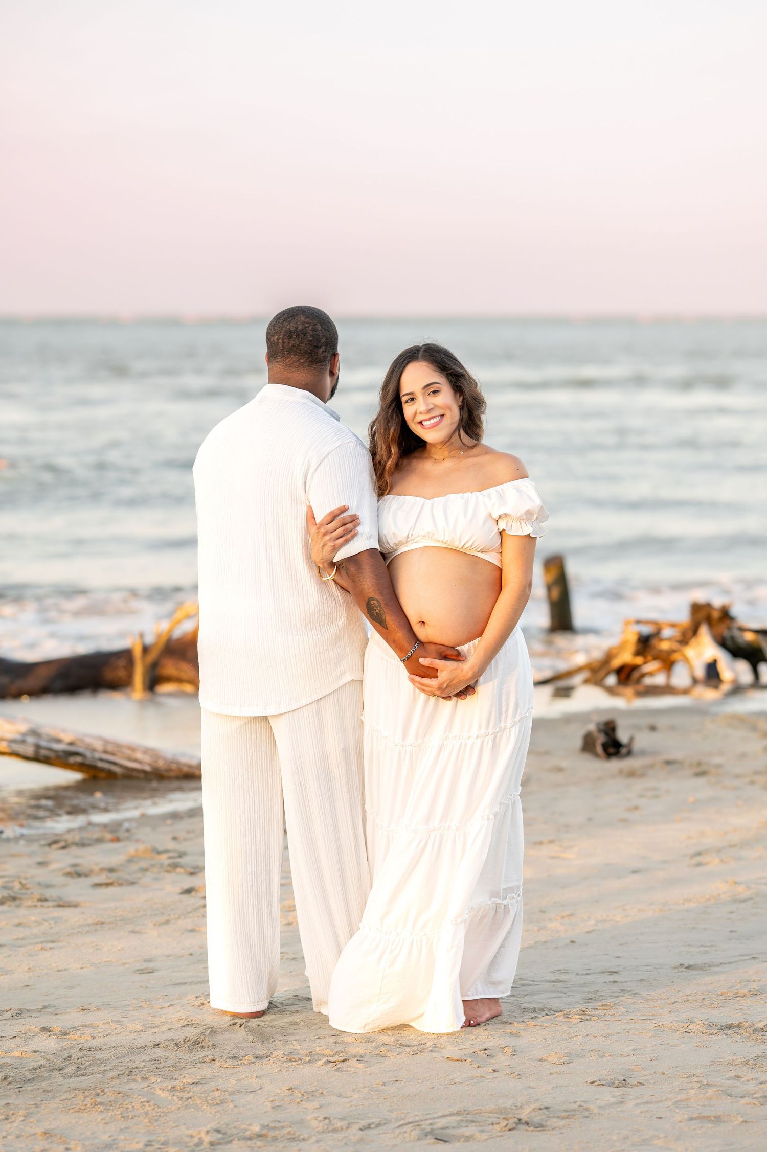 A couple in white outfits shares an intimate moment during a maternity photoshoot on the beach at sunset.