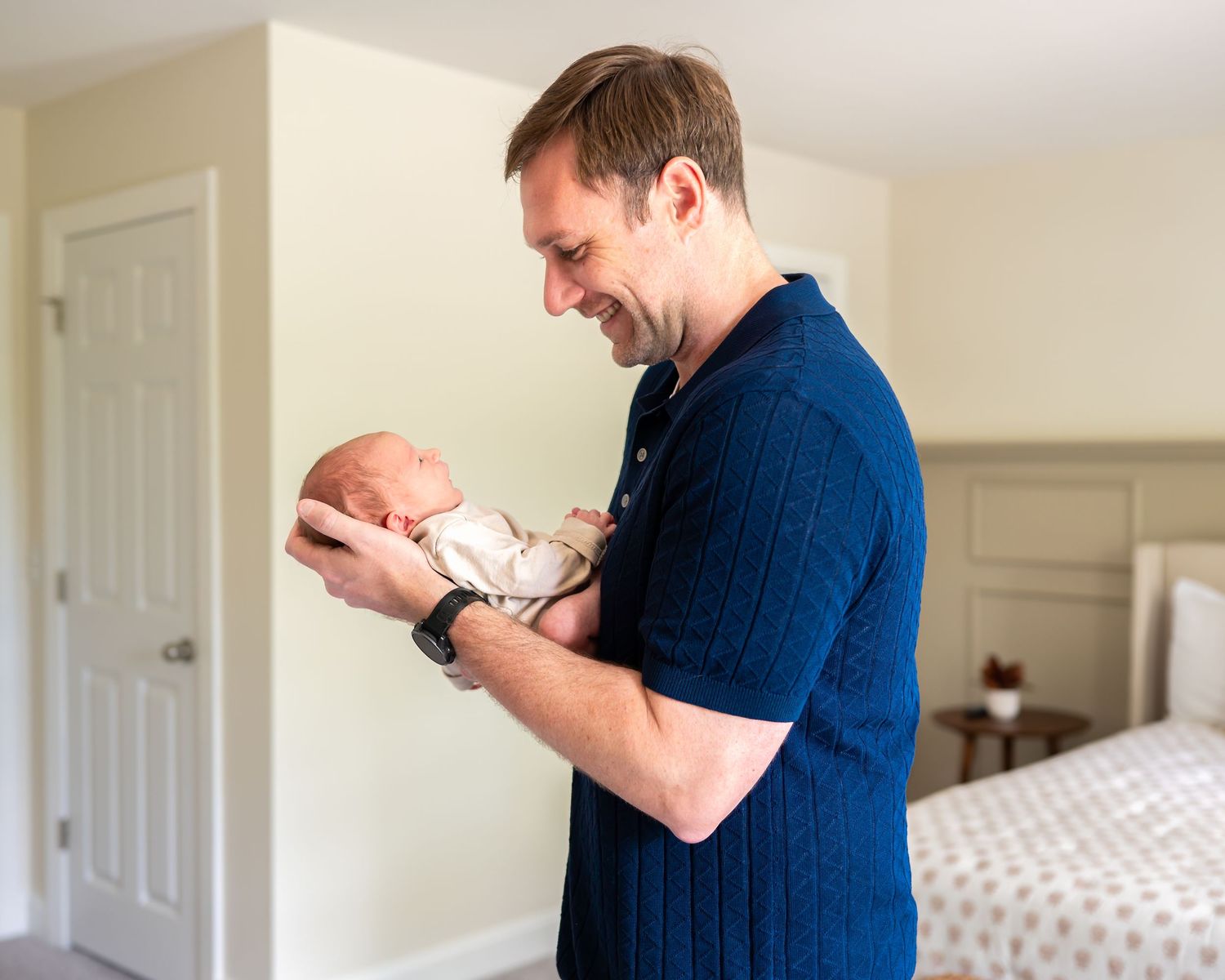 A smiling father in a navy blue polo shirt gently holds his newborn baby in a bright bedroom.