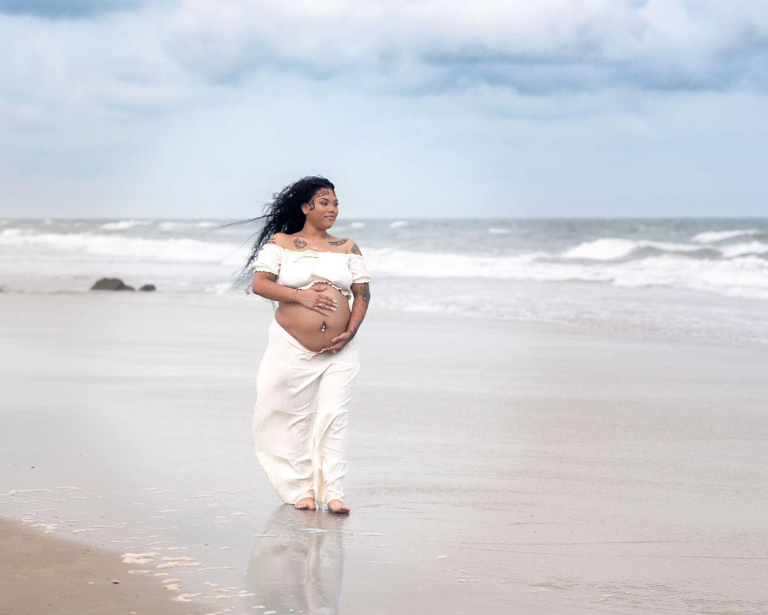 Pregnant woman in white dress standing on beach shore with ocean waves, holding baby bump, hair blowing in wind.