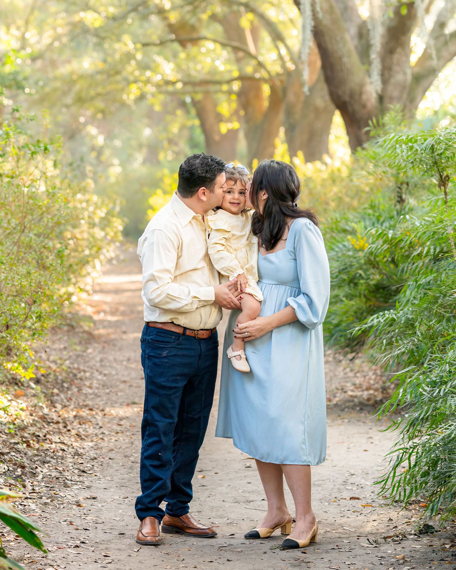 A family of three poses on a sunlit garden path, parents kissing their toddler while mom shows a baby bump in a blue dress.
