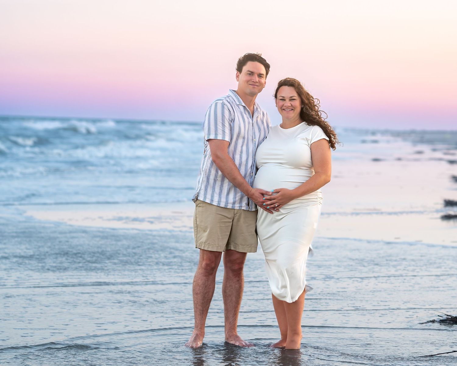 Couple poses on beach at sunset, woman in white dress cradling baby bump, waves and pink sky behind them.