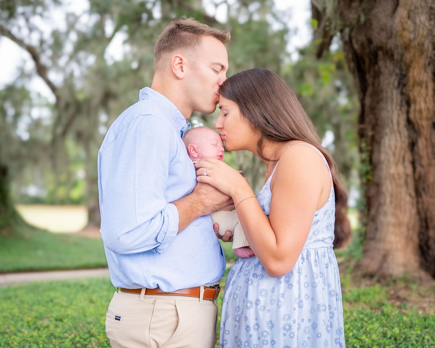 Mom and Dad both wearing light blue hold their newborn son between them and each kiss his head