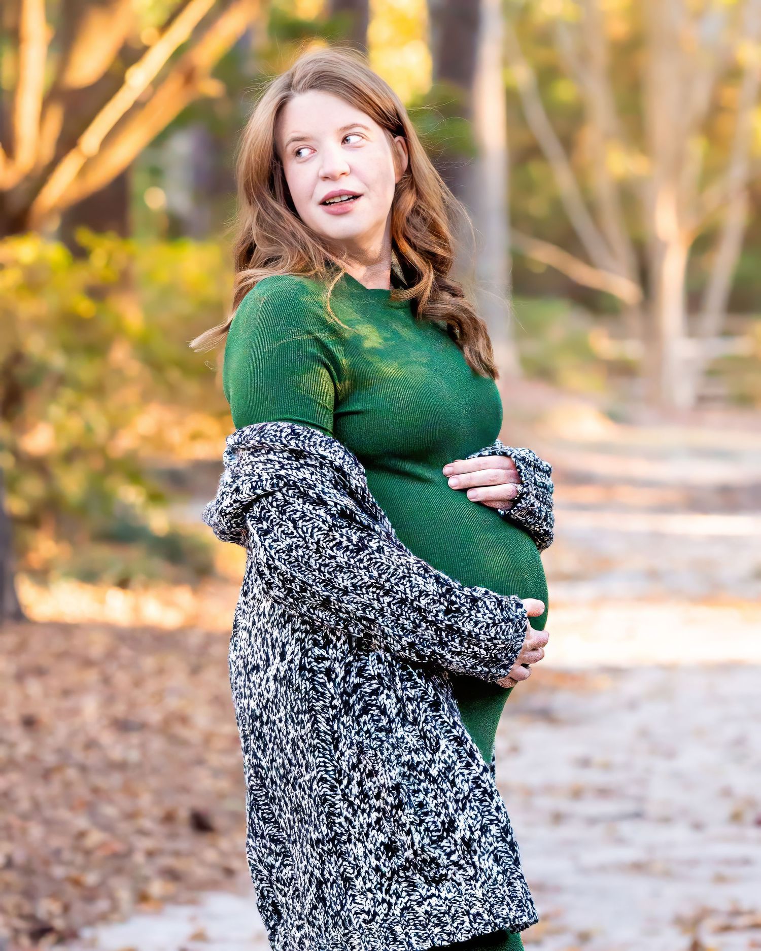 Person in green dress and cozy marled cardigan poses on path surrounded by fall foliage during autumn maternity session.