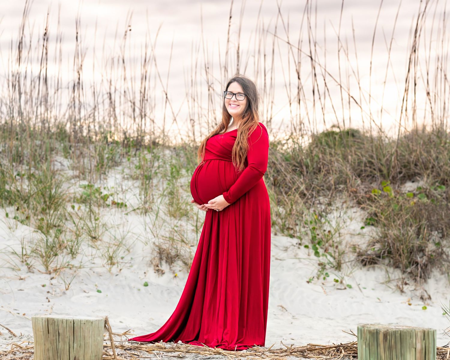 A pregnant woman in a flowing red maxi dress stands on a sandy beach among sea grass at sunset.