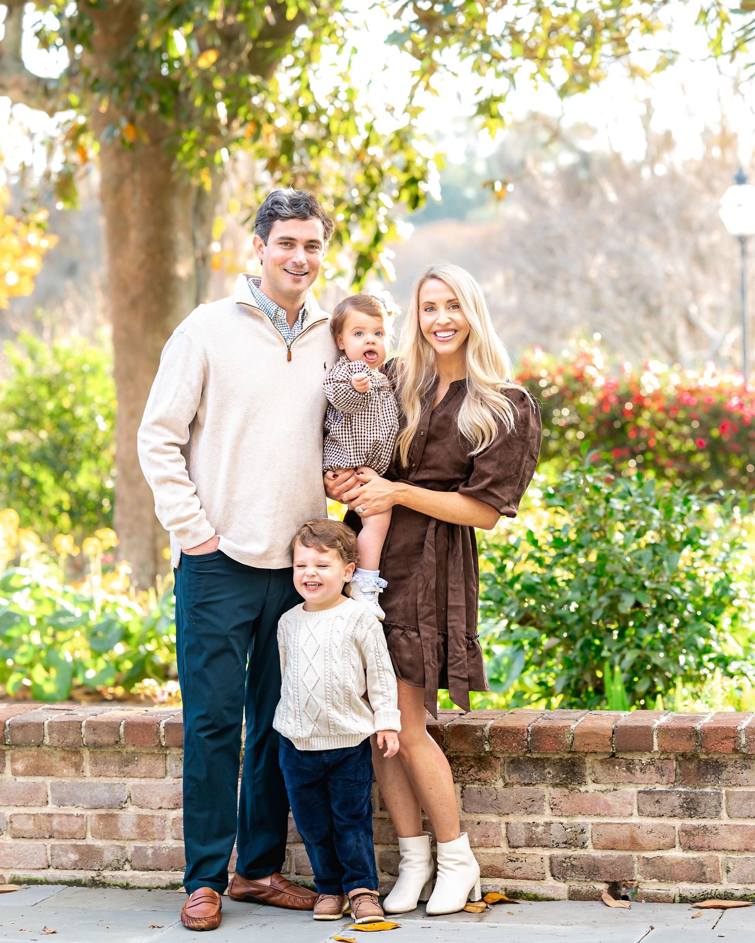 Young family of four posing outdoors in fall attire, parents with two toddlers in front of brick wall and green trees.
