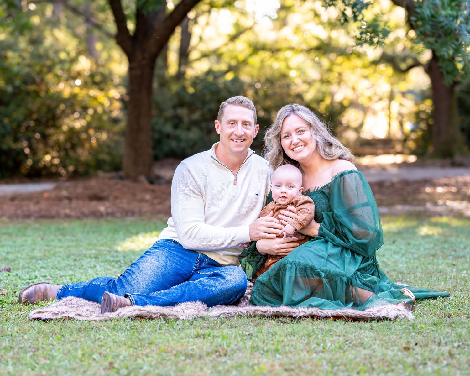 Family relaxing together on a blanket in a sunny park setting.