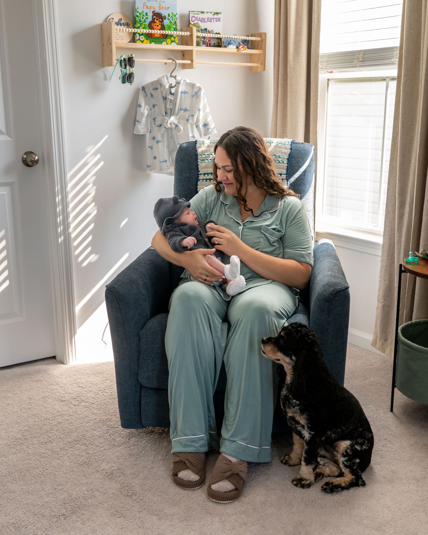 A mother in green pajamas bottle-feeds her newborn in a blue nursery chair while a black dog sits beside her.