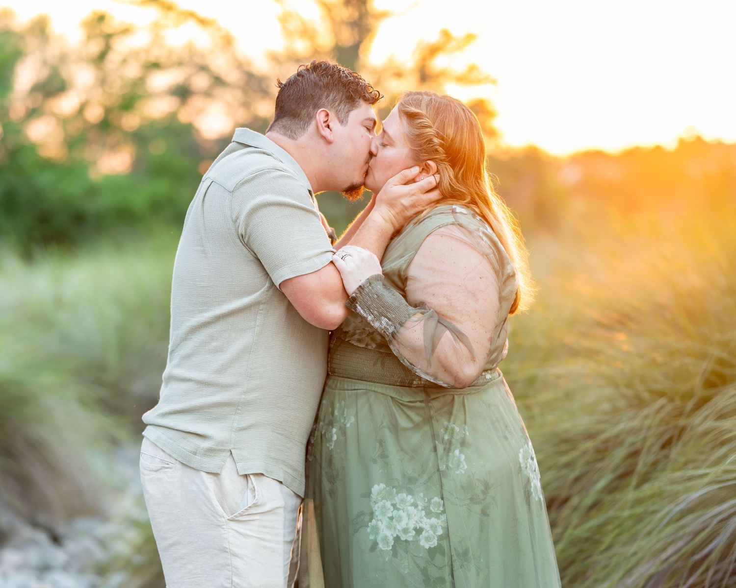 Couple sharing a romantic kiss at golden hour in a lush green field, woman in floral green dress.