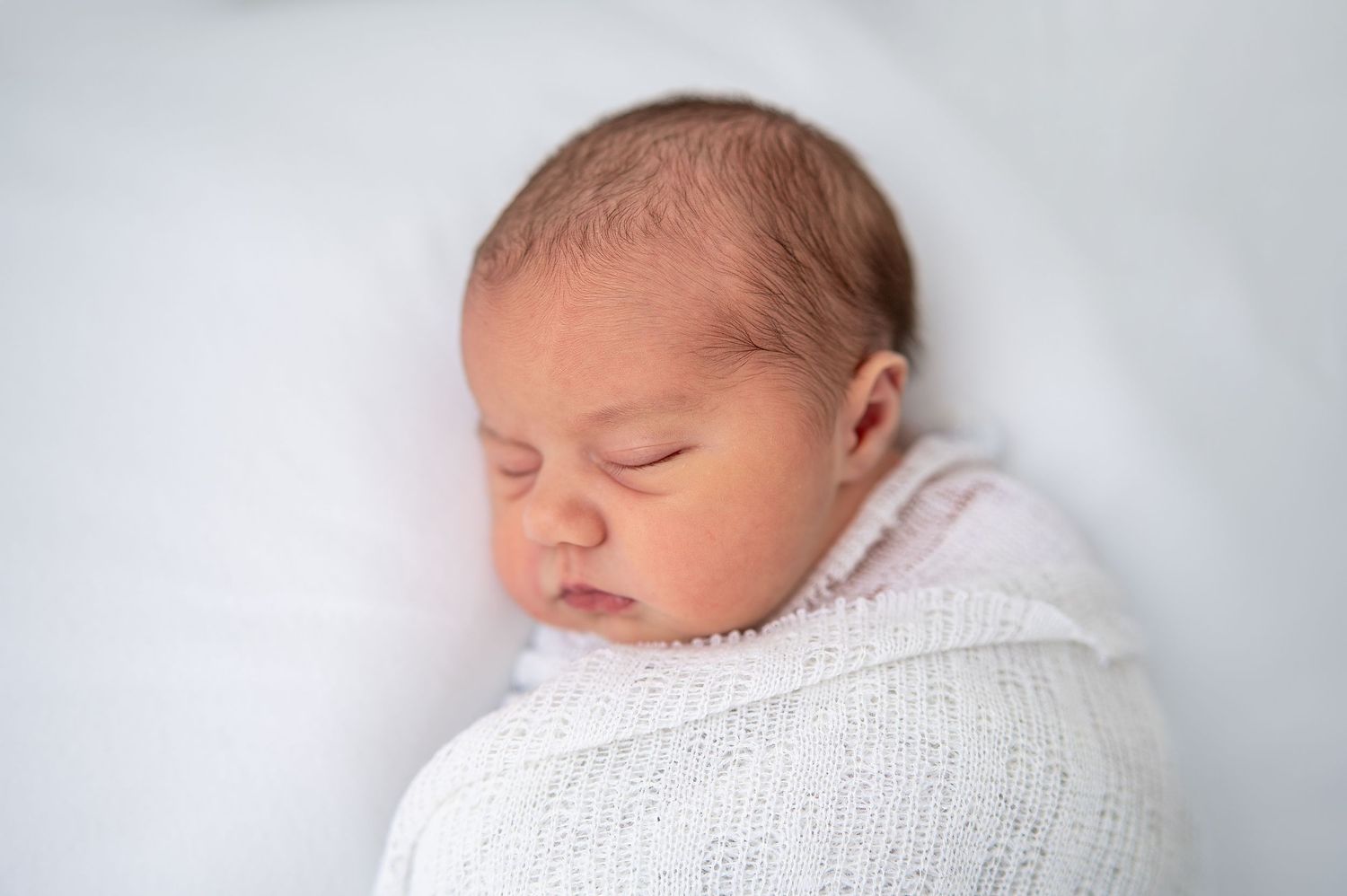 Peaceful newborn baby sleeping swaddled in a soft white knit wrap on a white background.