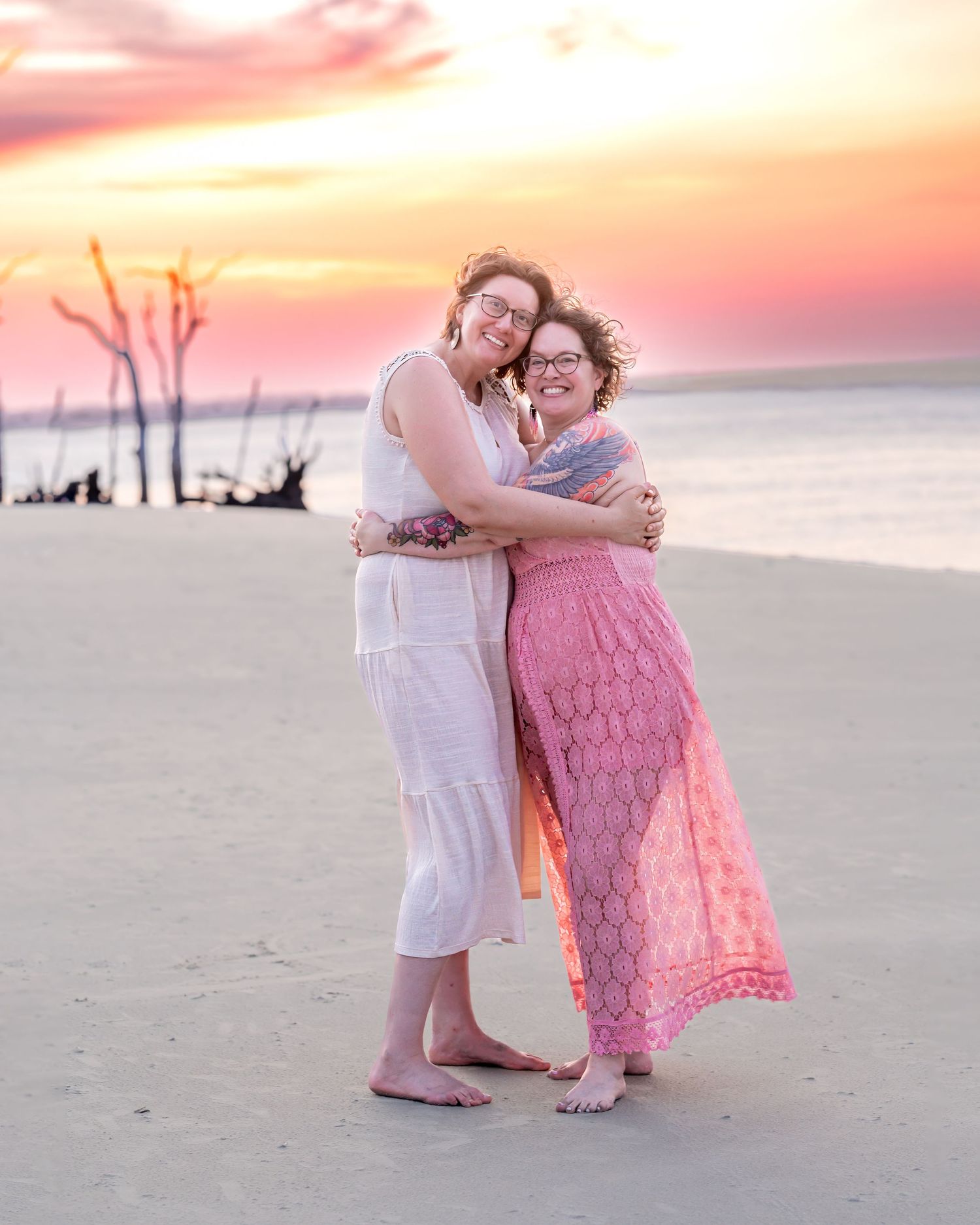 Two women hugging and smiling on a sandy beach at sunset with pink and orange skies in the background.