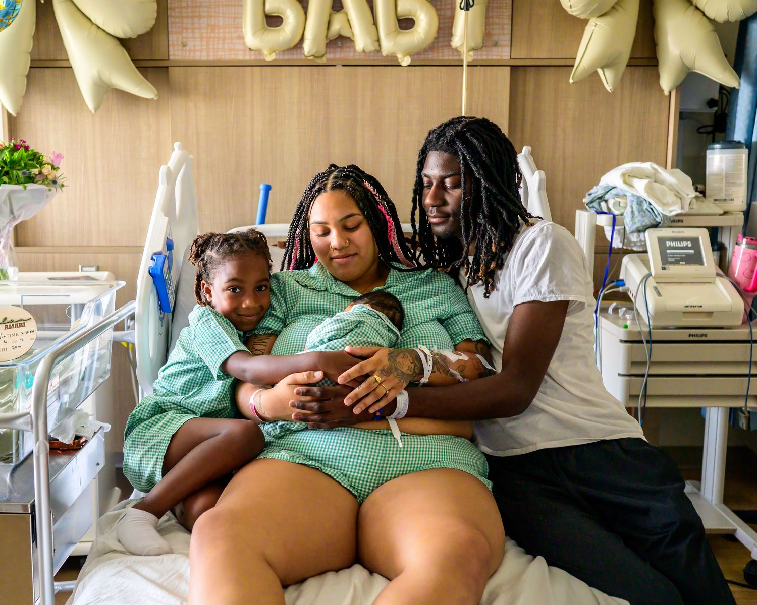 Family in hospital room, parents holding newborn baby with toddler sibling, celebrating new arrival under BABY balloon.