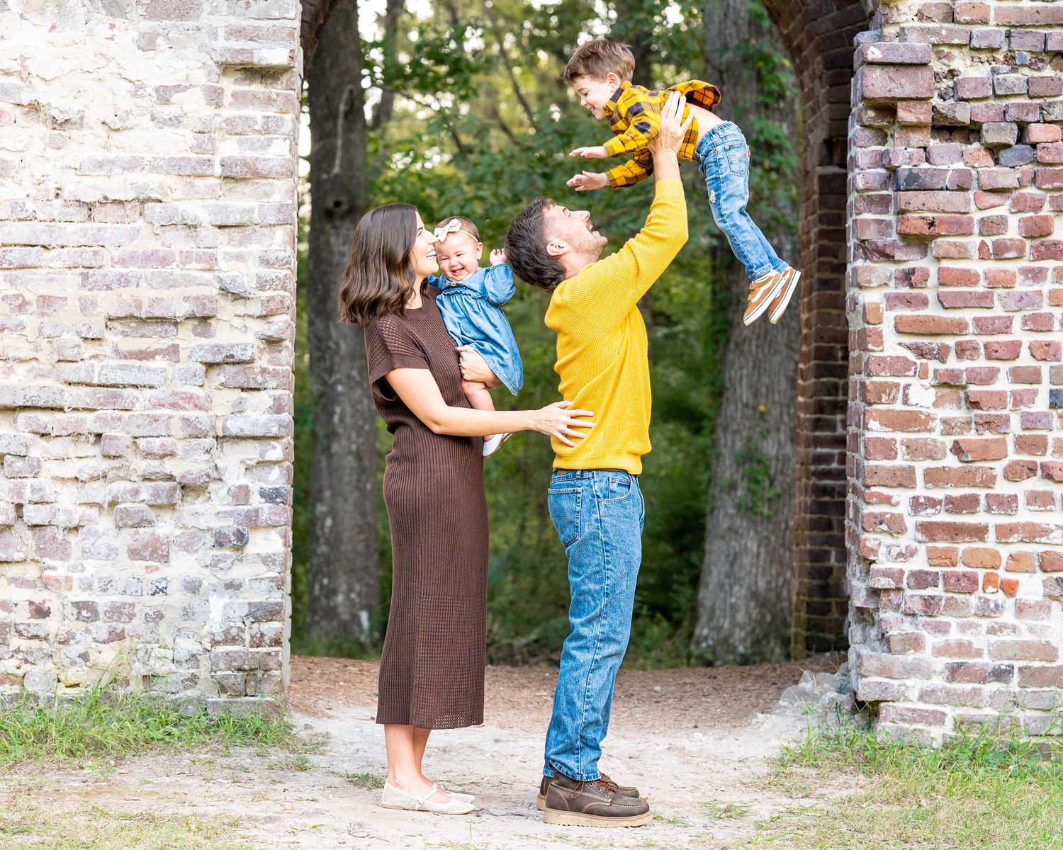 Family of four playing outdoors near rustic brick archway, parents lifting children in autumn setting.