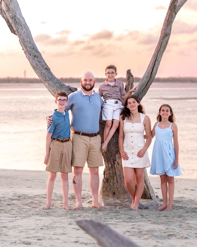 Family of five posing by a beach tree at sunset, wearing coordinated light blue and khaki outfits on sandy shore.