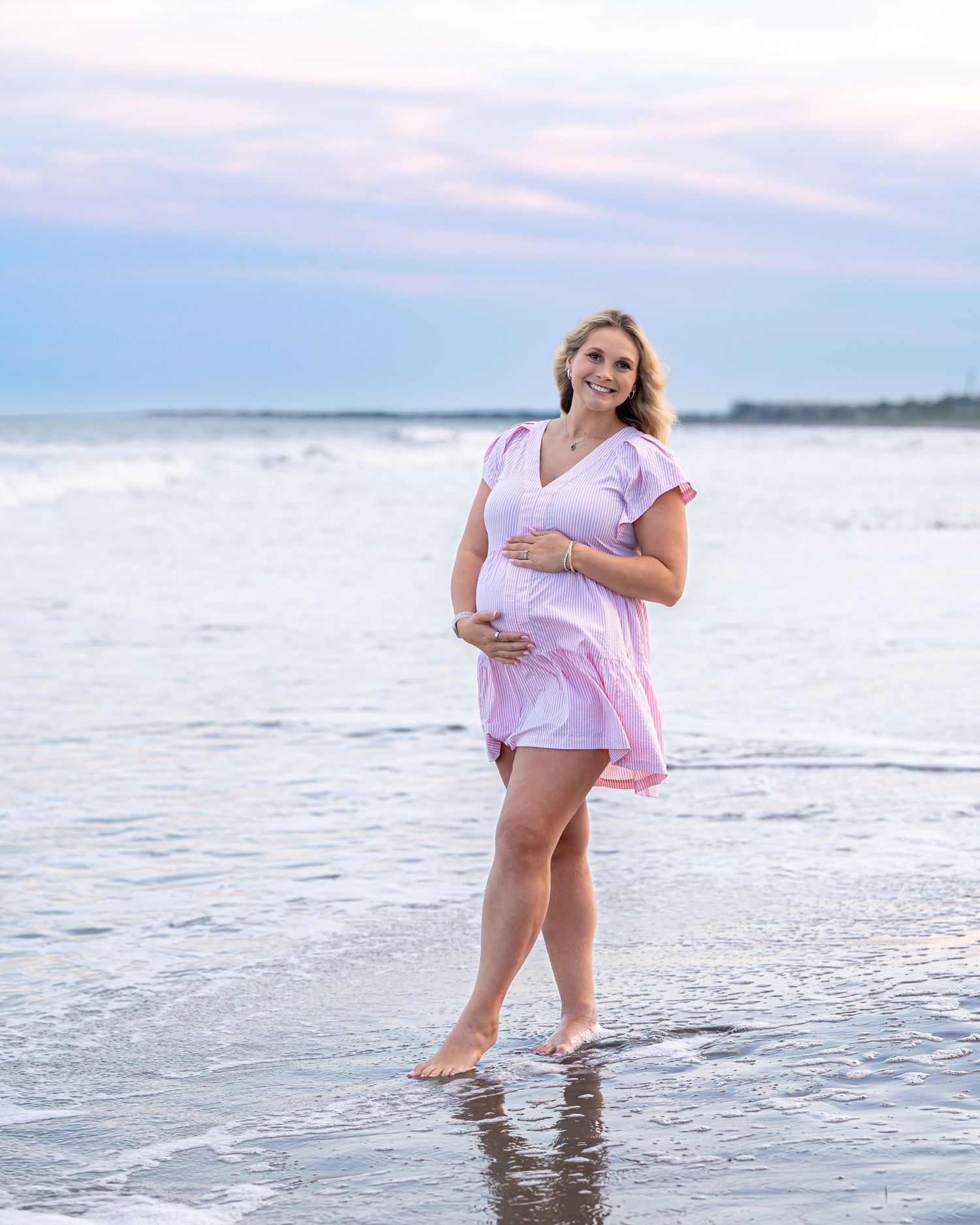 Pregnant woman in pink dress standing barefoot in ocean waves at sunset during a maternity beach photo session.