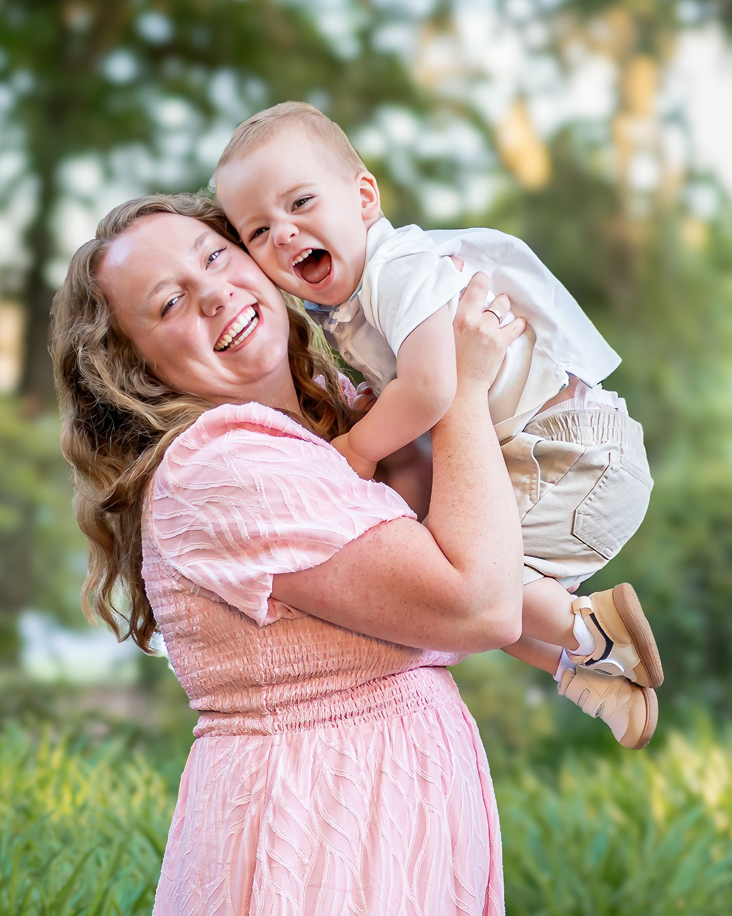 A joyful mother in a pink dress lifts her laughing toddler boy outdoors in a lush green park setting.