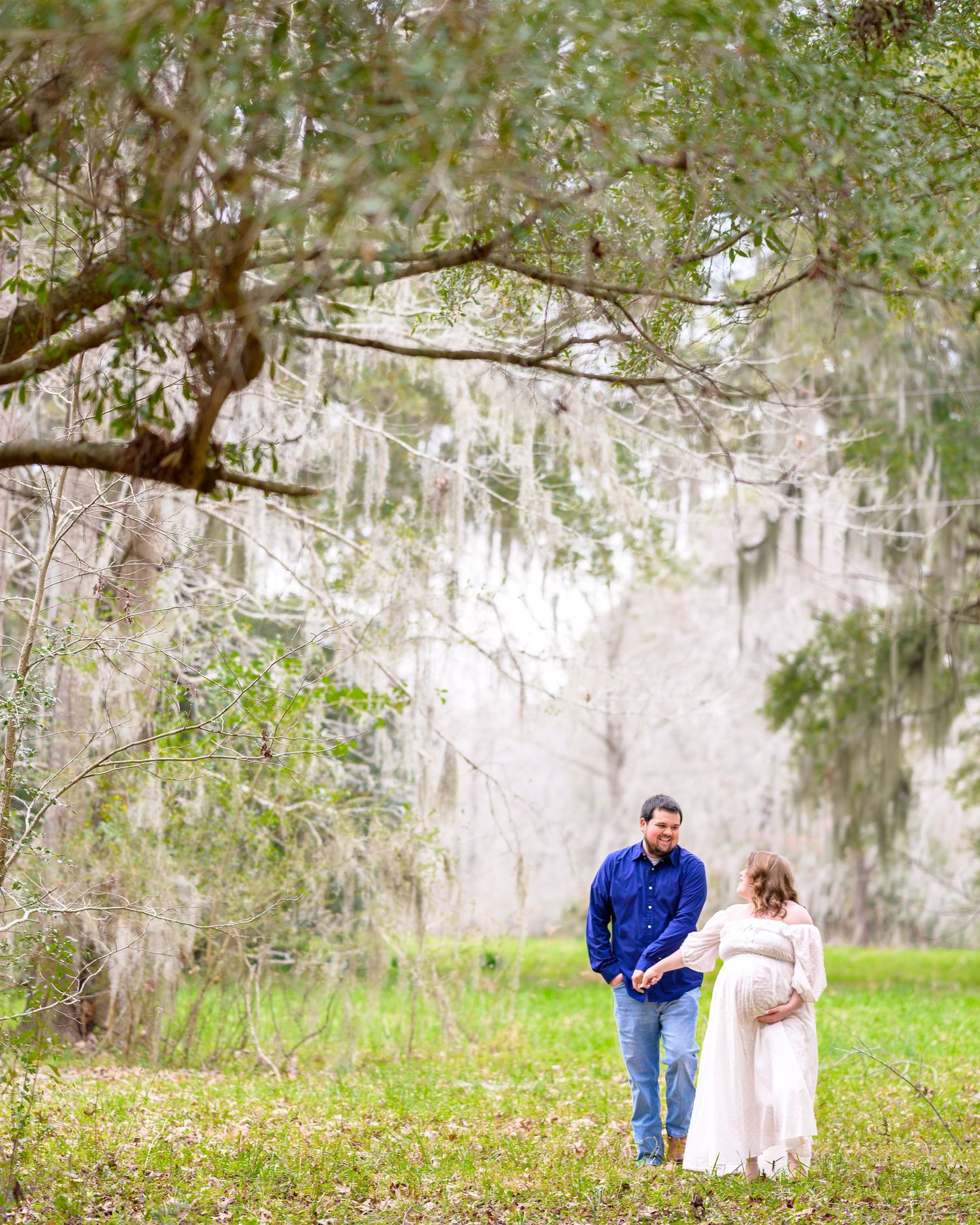 Erin and her husband walk hand in hand under a large tree in a wide open field during their maternity session.