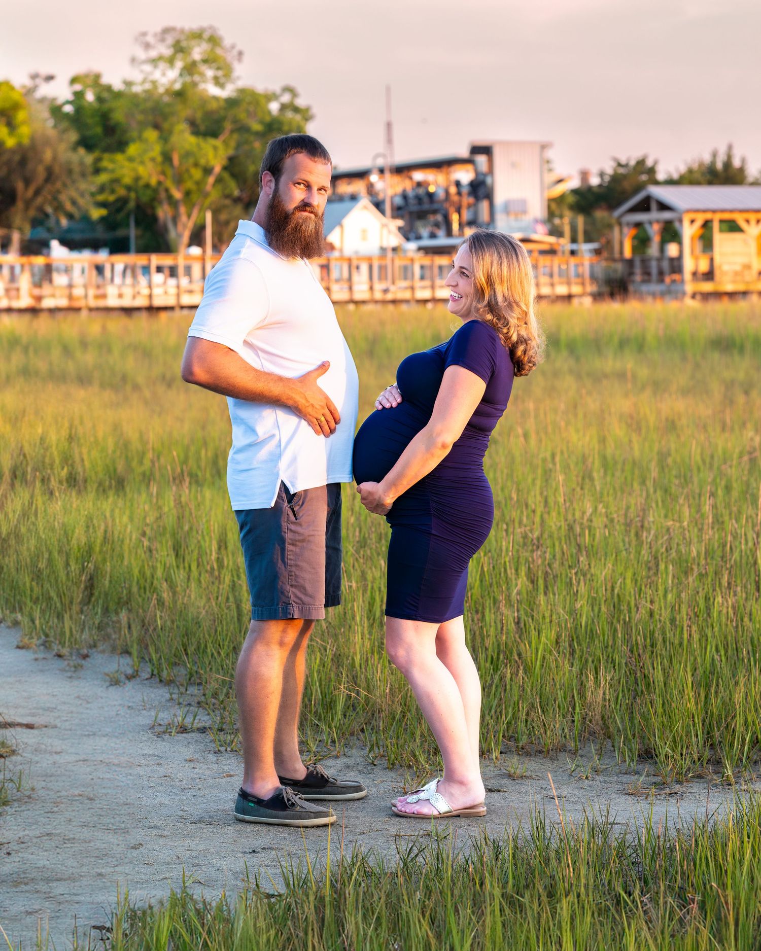 A couple stands together on a marsh path during golden hour with coastal buildings in the background.