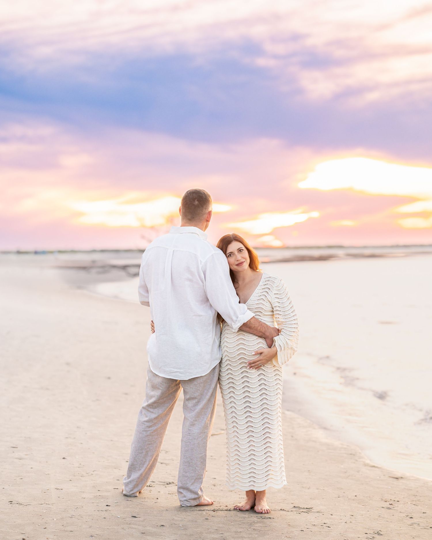 Pregnant couple embracing on beach at sunset, woman in white lace maternity gown cradling bump, colorful pastel sky backdrop.