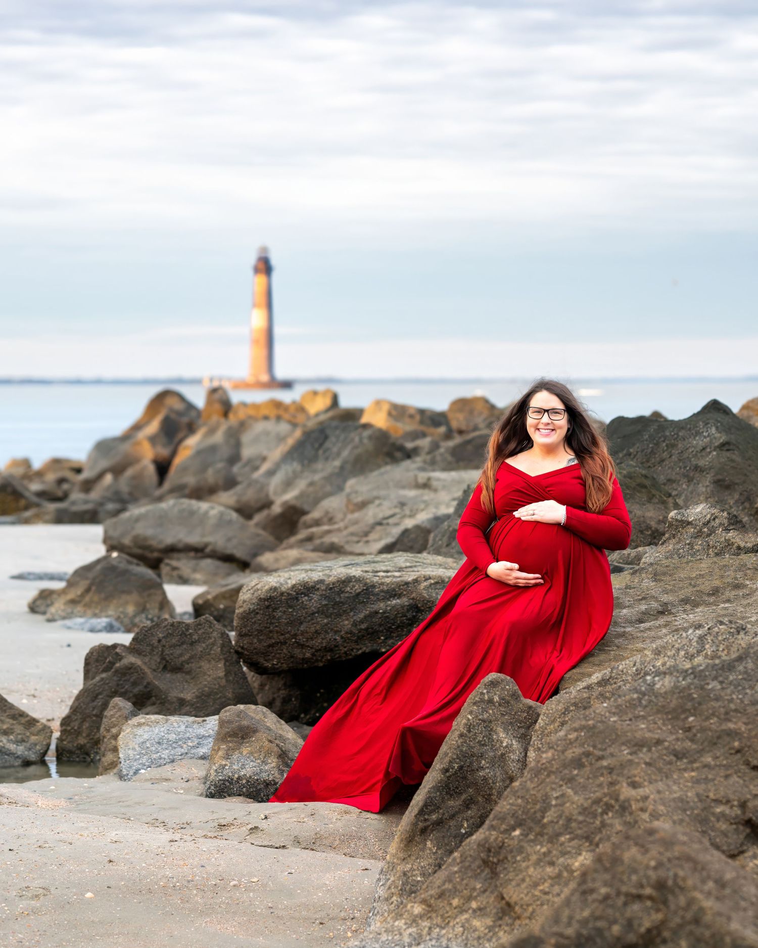 Pregnant woman in a flowing red dress poses gracefully on rocky coastal shoreline with a lighthouse in the background.