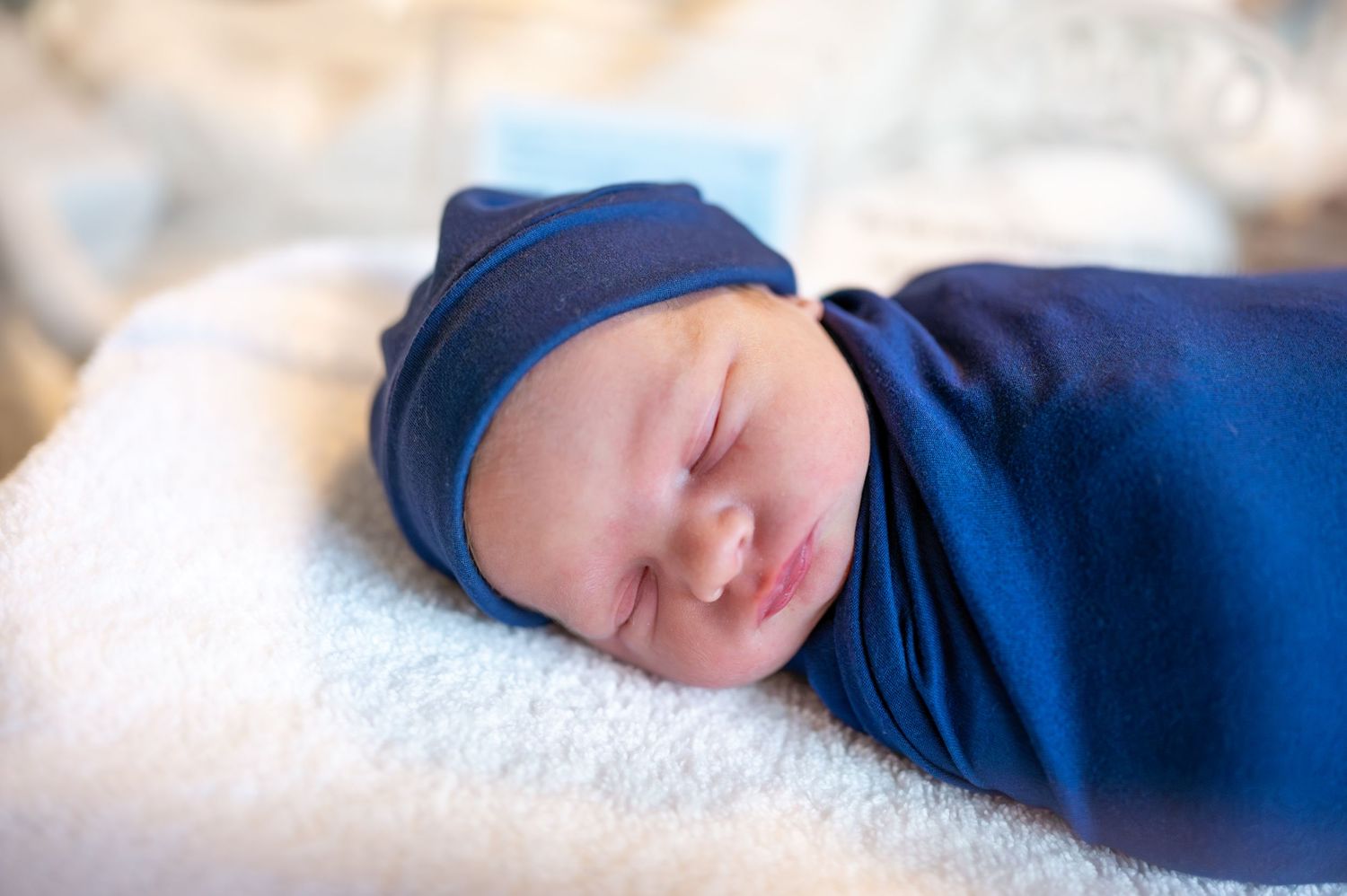 Peaceful sleeping newborn wearing navy blue hat and swaddle on white blanket.