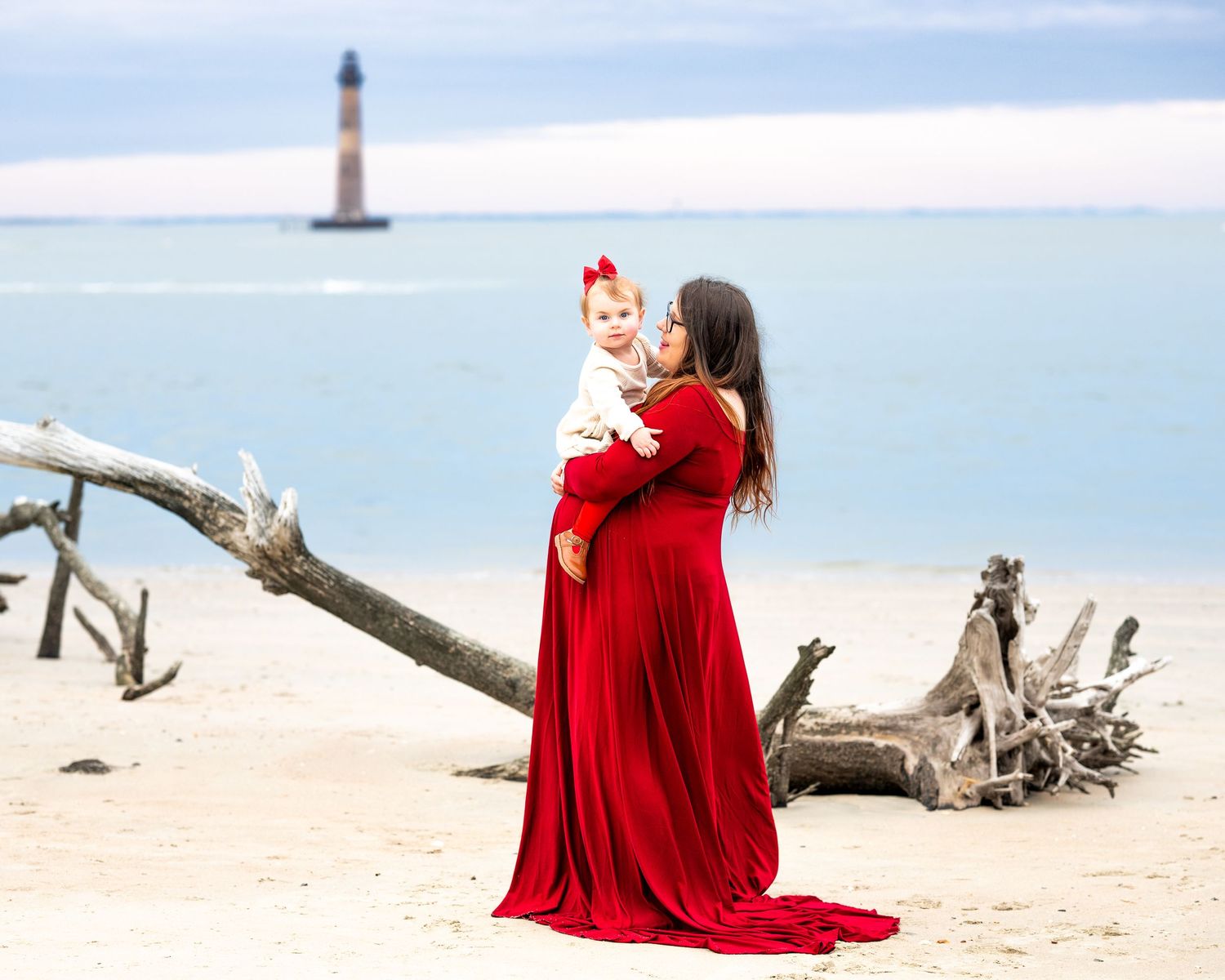 Woman in flowing red dress stands on beach with lighthouse visible in background over ocean waters.
