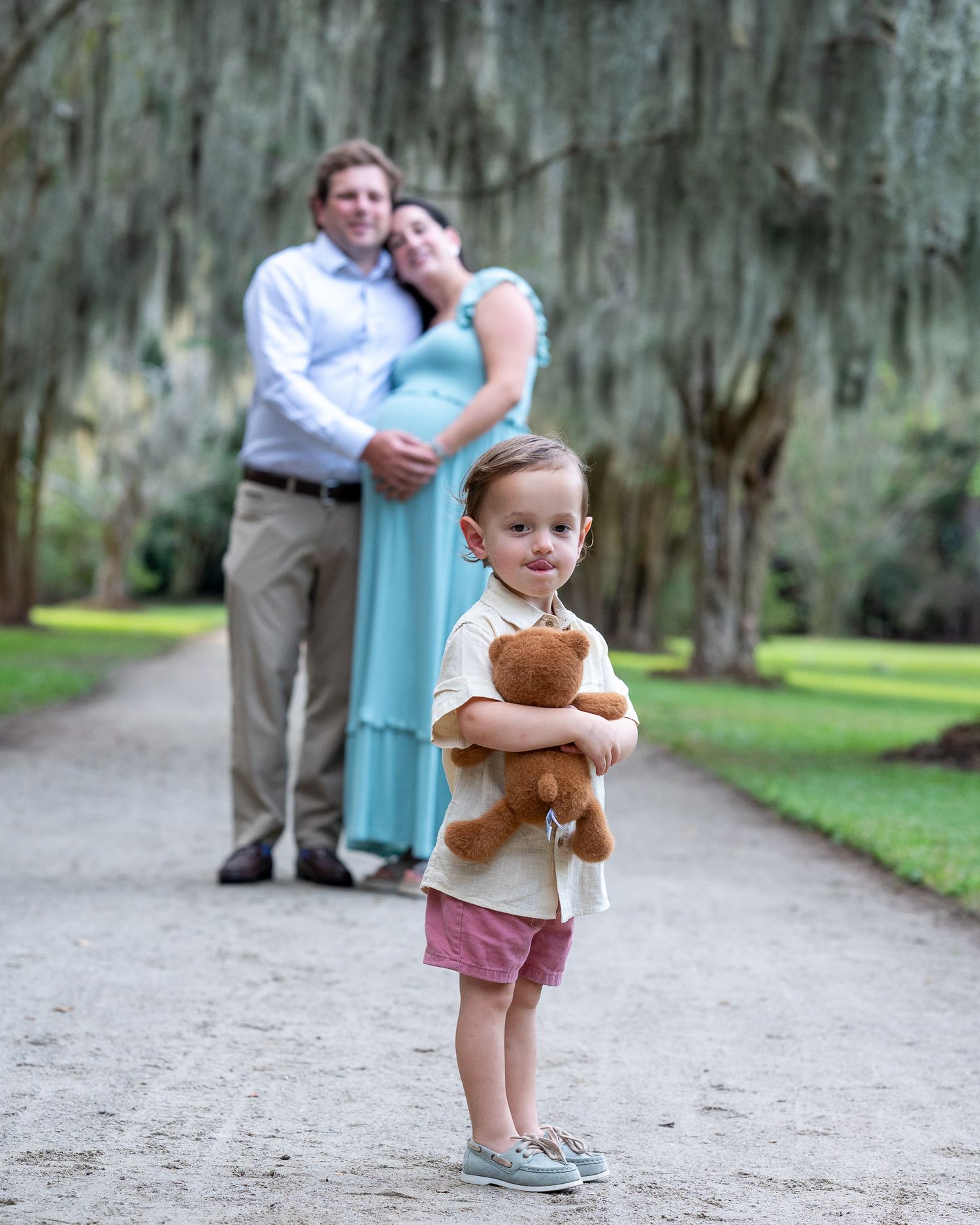 A a small child playing with a teddy bear along a tree-lined path while adults watch in the background.