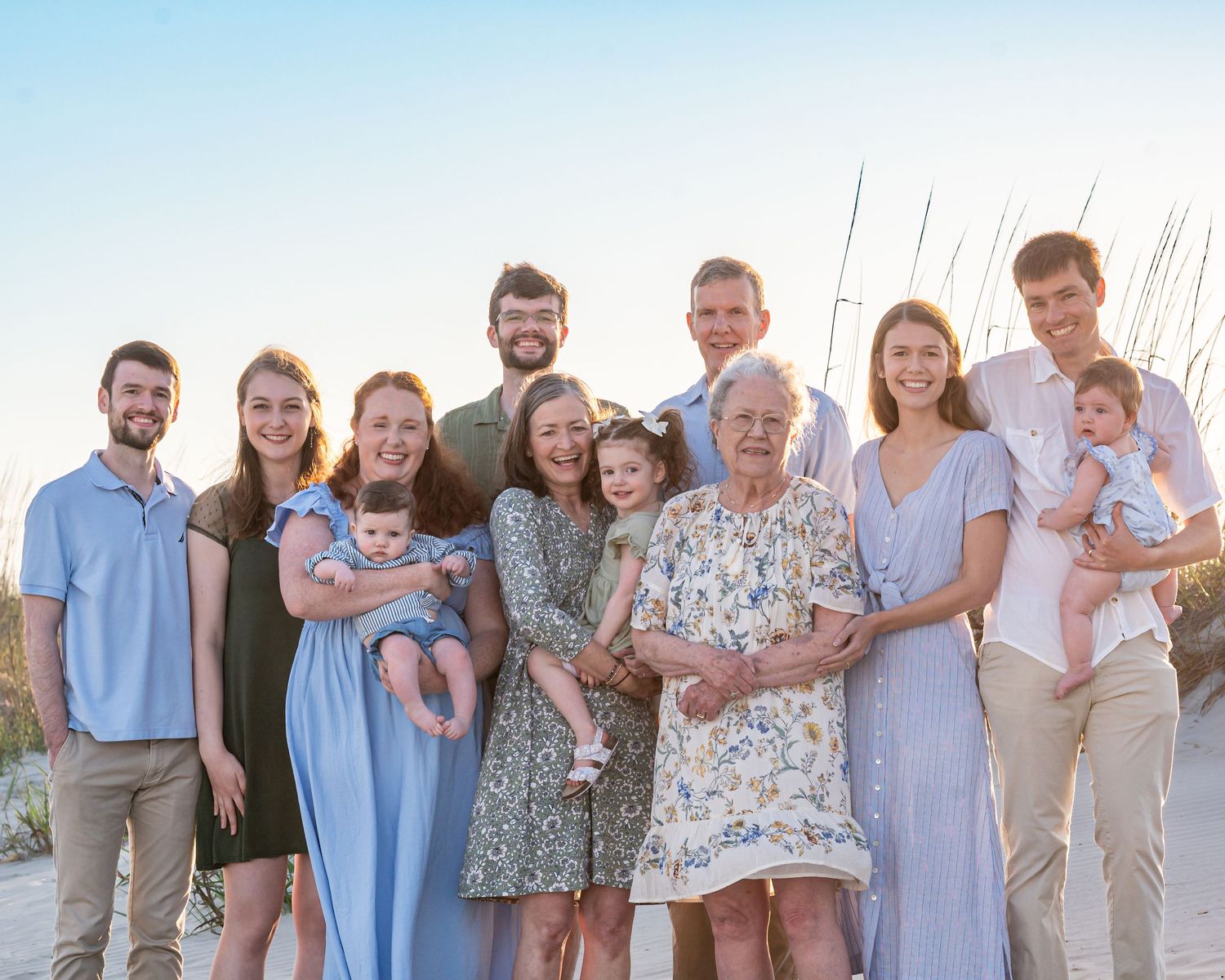 Large multi-generational family posing together on a beach at sunset, wearing coordinated blue and floral outfits.