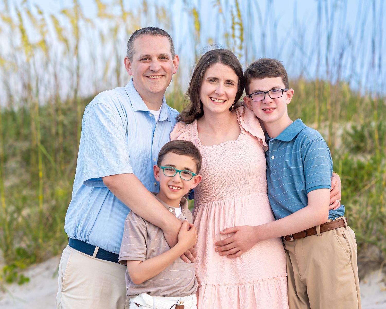 Family of four smiling together on a sandy beach with sea oats, two boys wearing glasses, parents embracing them warmly.