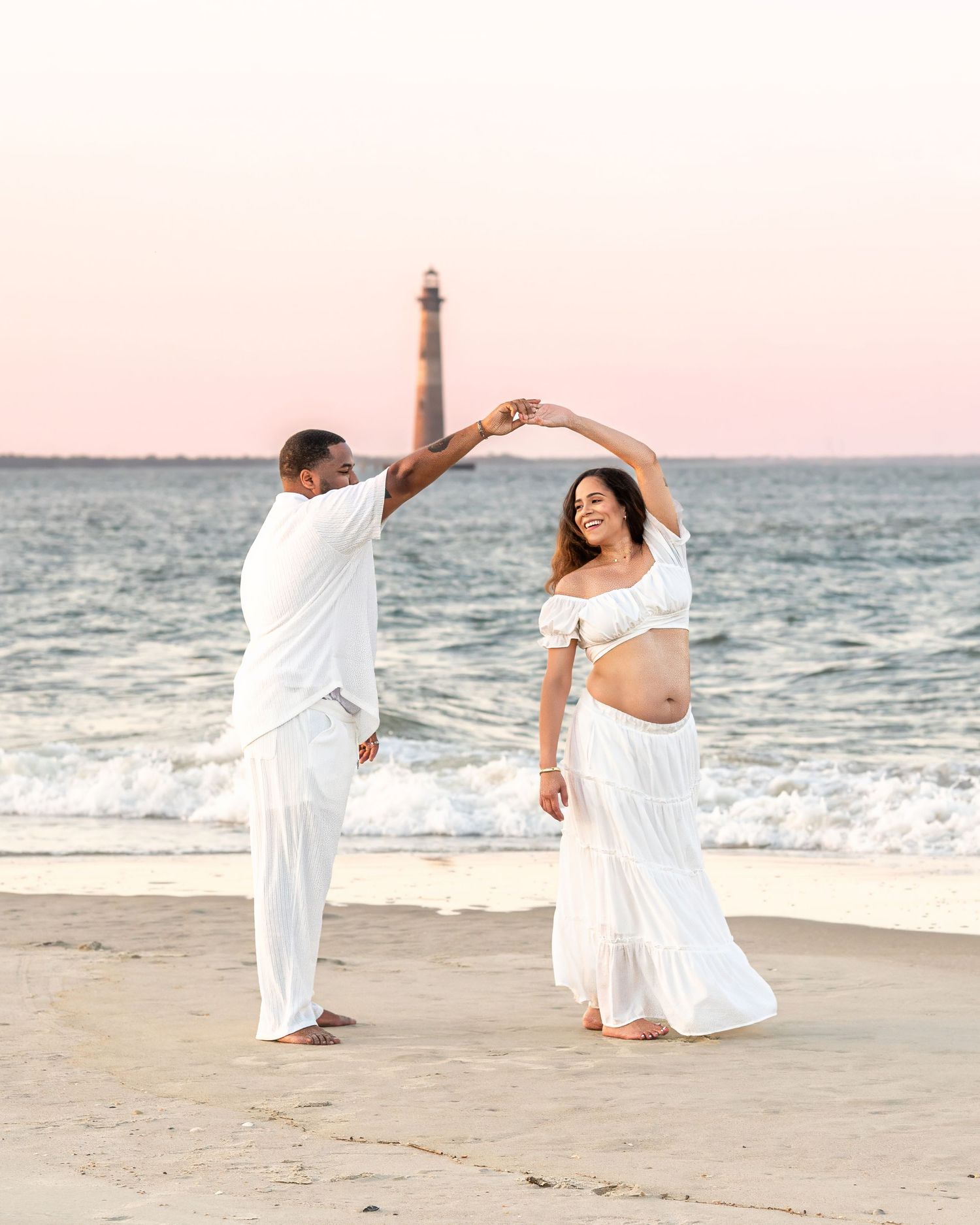 A couple in white clothing dances on the beach with a lighthouse visible in the distance.