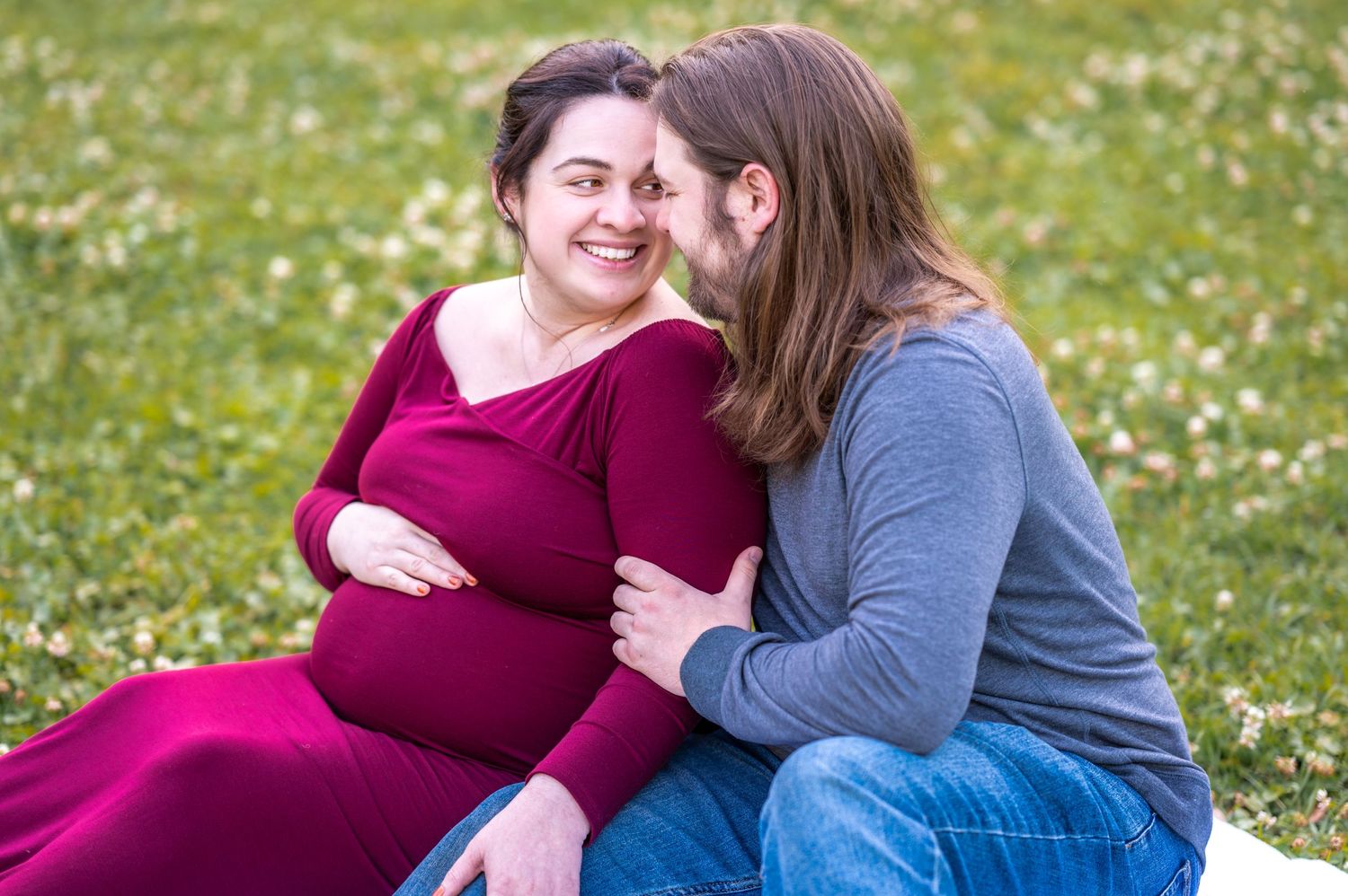 A couple sits together in a grassy field with white wildflowers blooming on a sunny day.