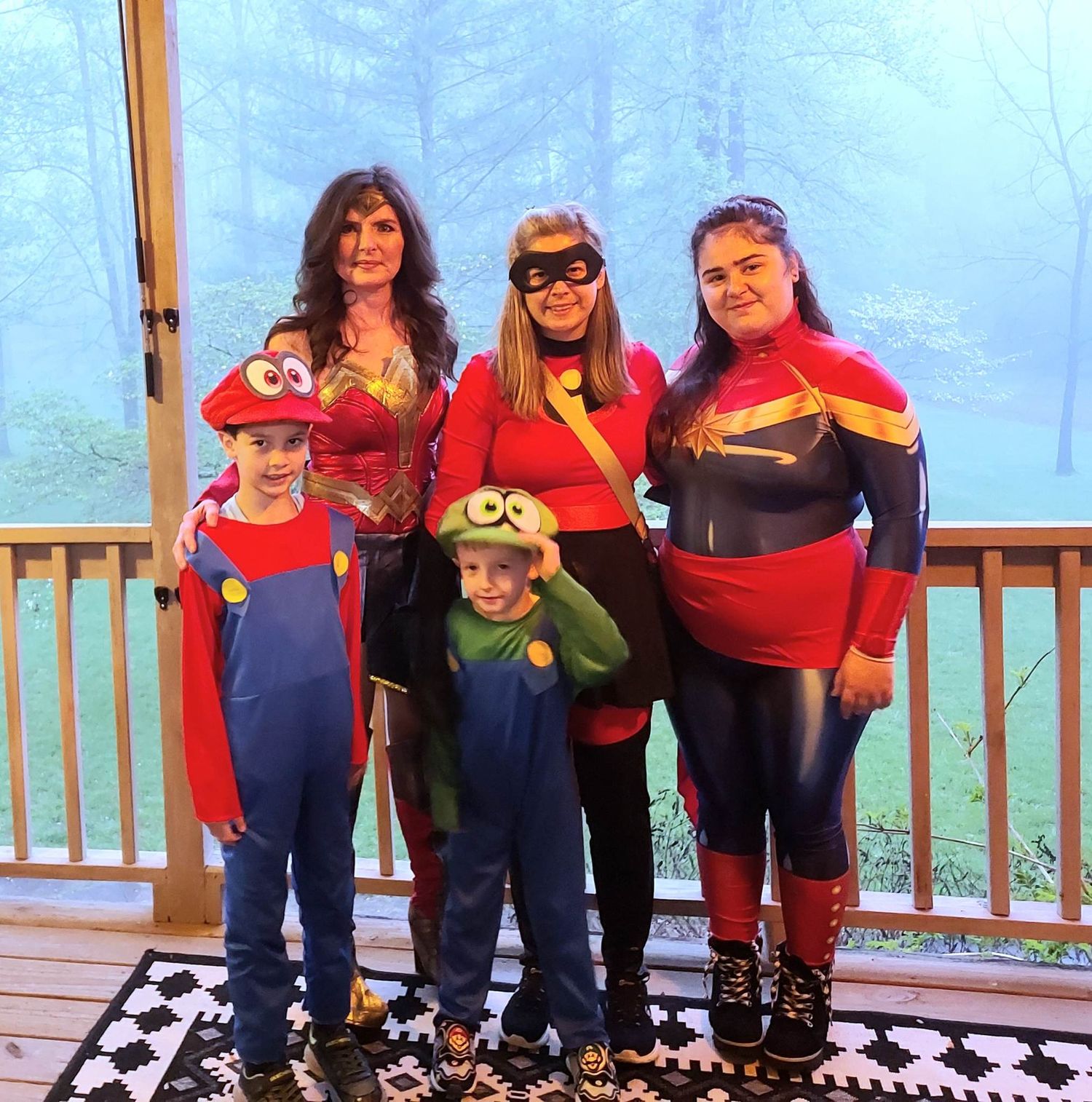A cropped and edited version of three adults dressed in superhero costumes and two children dressed as Mario and Luigi pose for a photo