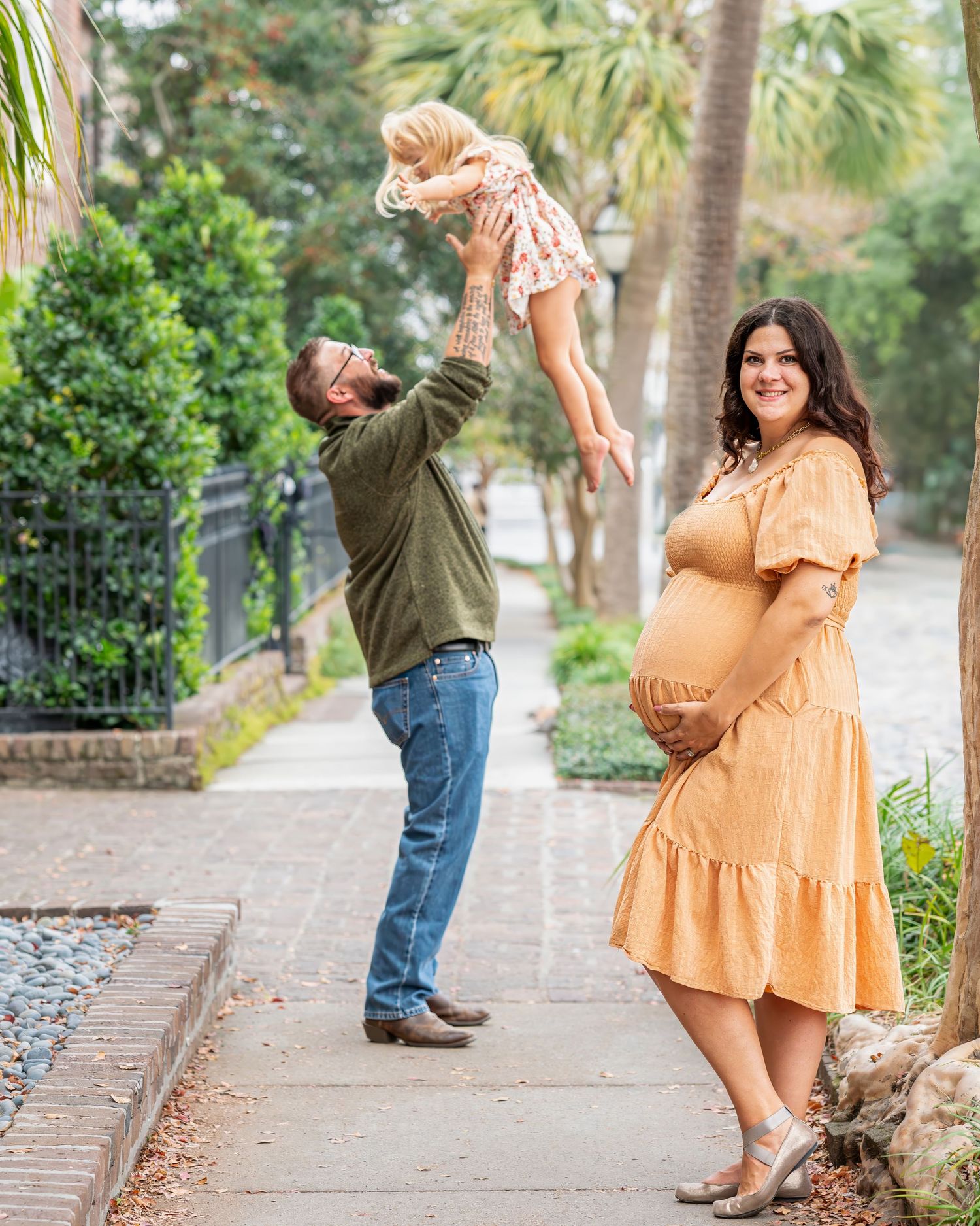 Josie stands on a sidewalk as her husband is behind her throwing their laughing toddler into the air and catching her.