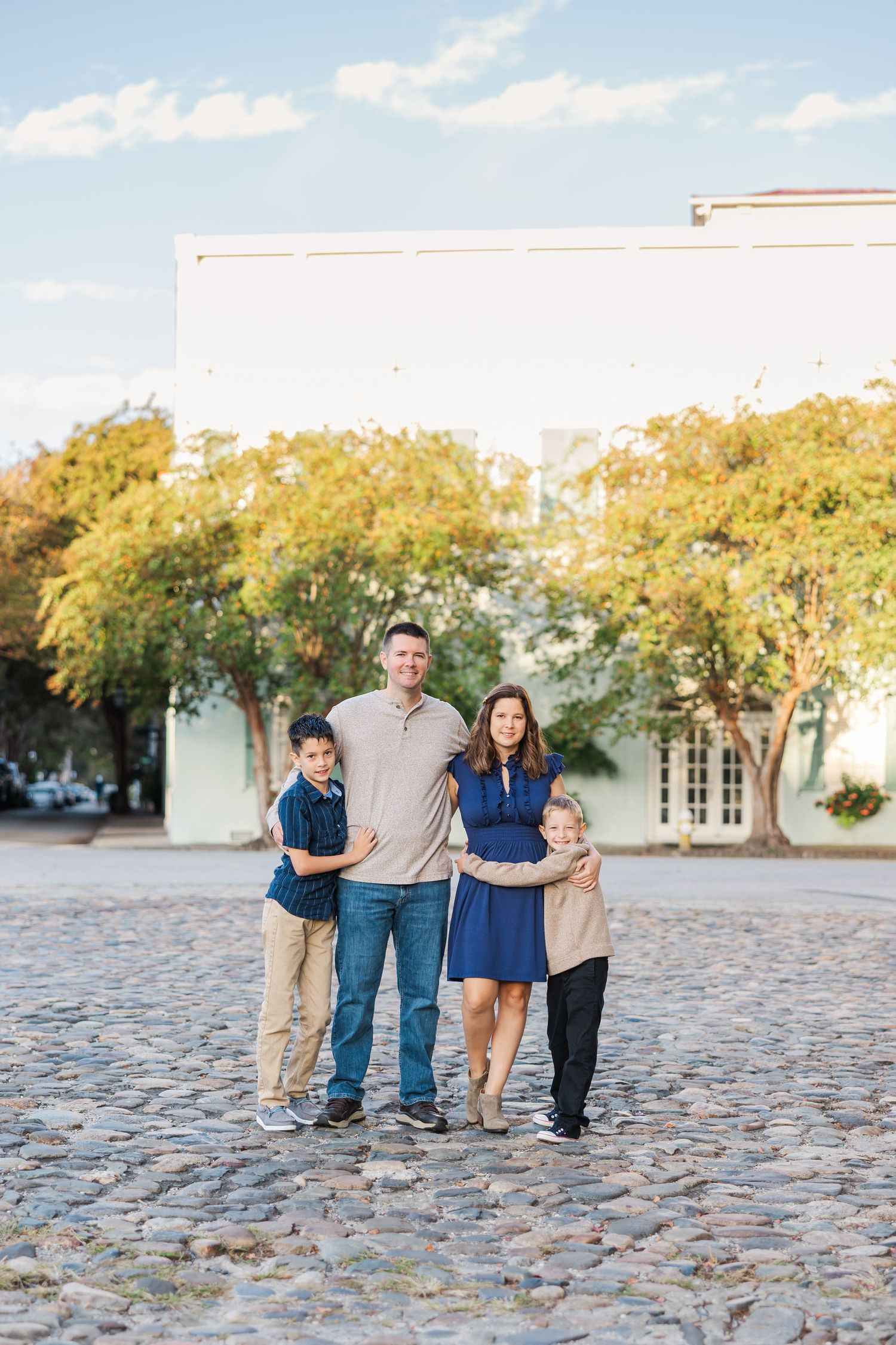 Family walking together on cobblestone path surrounded by autumn trees and white building in background.